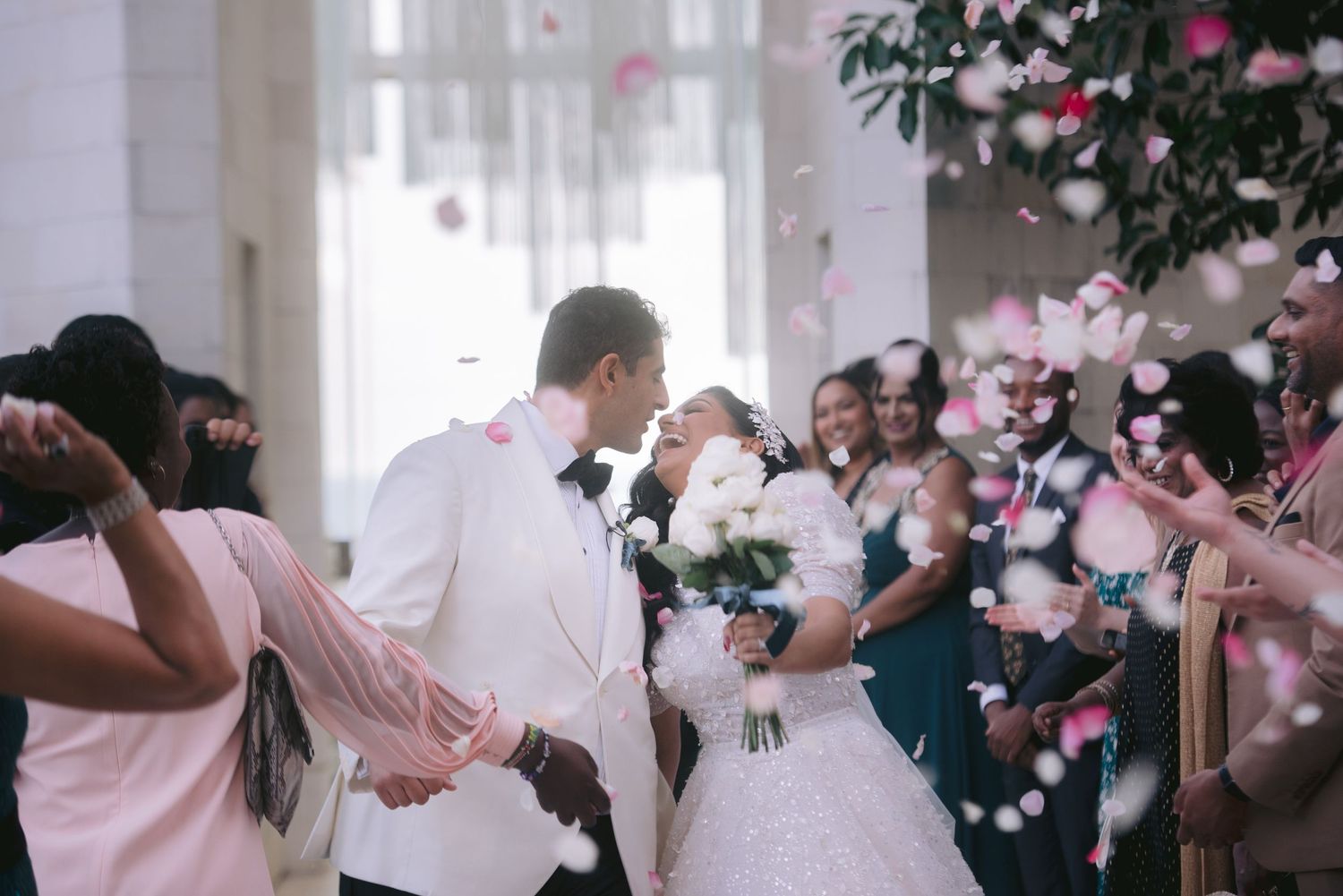 Newlywed couple sharing a kiss while guests toss pink rose petals during a joyful wedding ceremony.