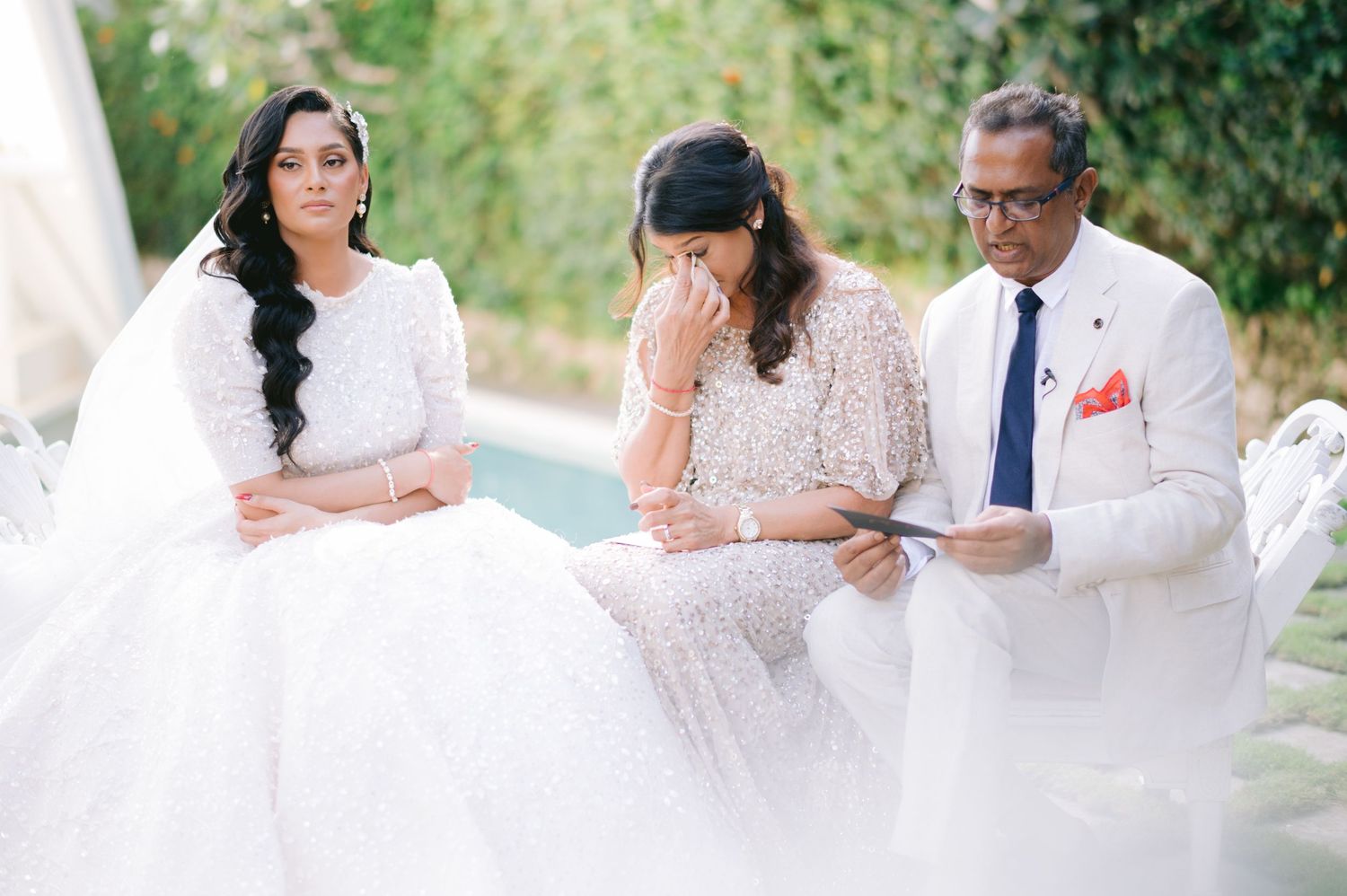 Emotional wedding ceremony with bride in white gown, tearful woman in beaded dress, and officiant reading vows outdoors.