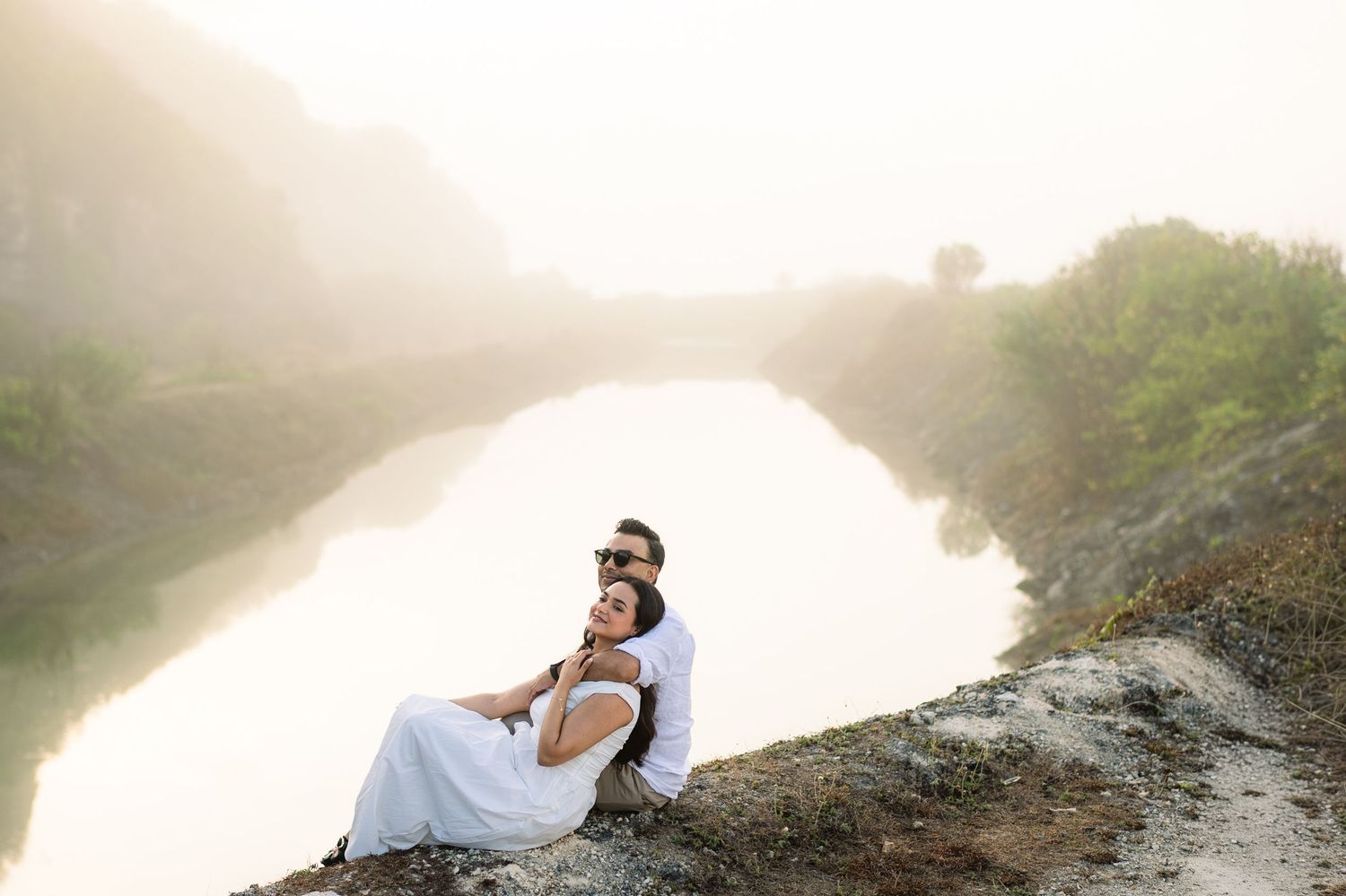 Couple embracing on rocky hillside with serene misty river backdrop in soft golden light.