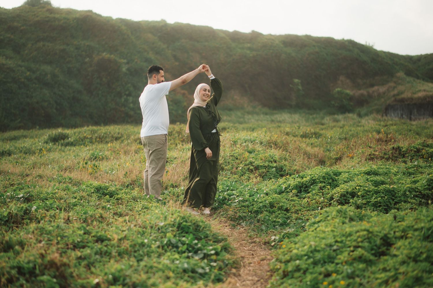 A couple dances together in a lush green meadow, the man spinning the woman in a hijab along a winding path.