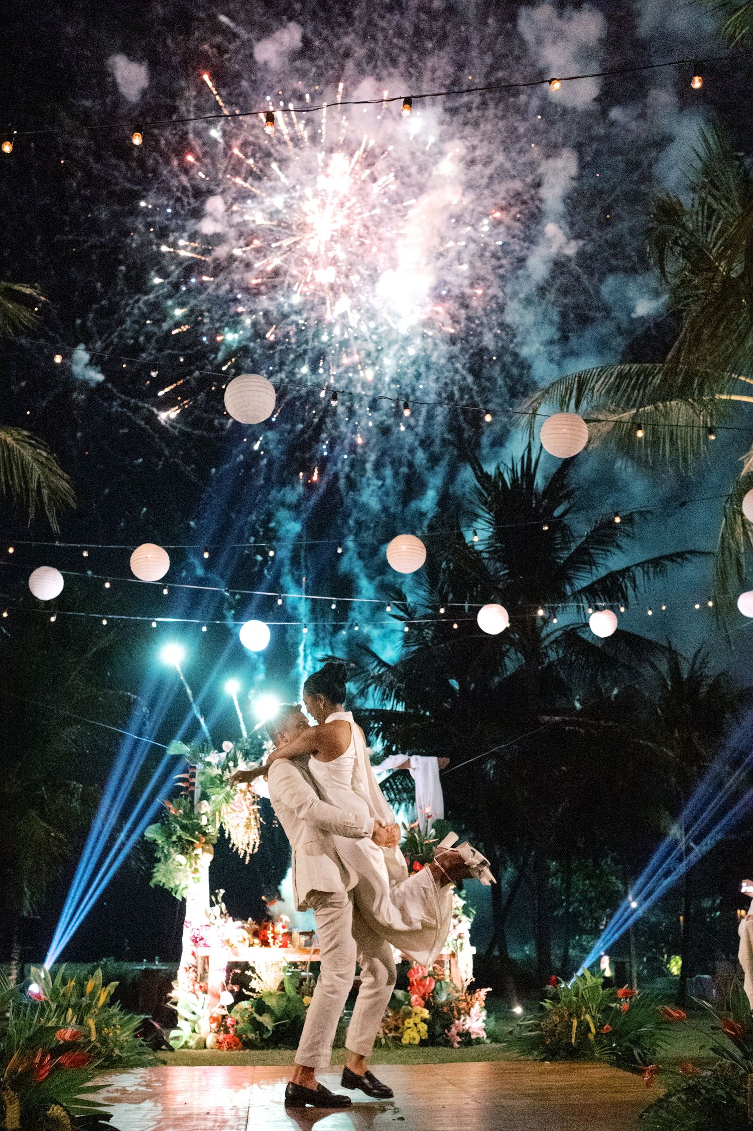 Couple sharing first dance lift under spectacular fireworks display with string lights and palm trees at tropical wedding.