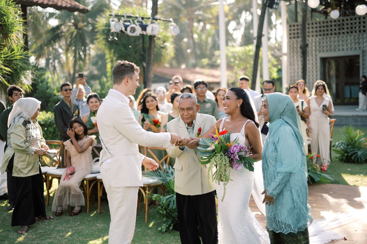 Outdoor wedding ceremony with couple receiving traditional blessing from elder guests in tropical setting.