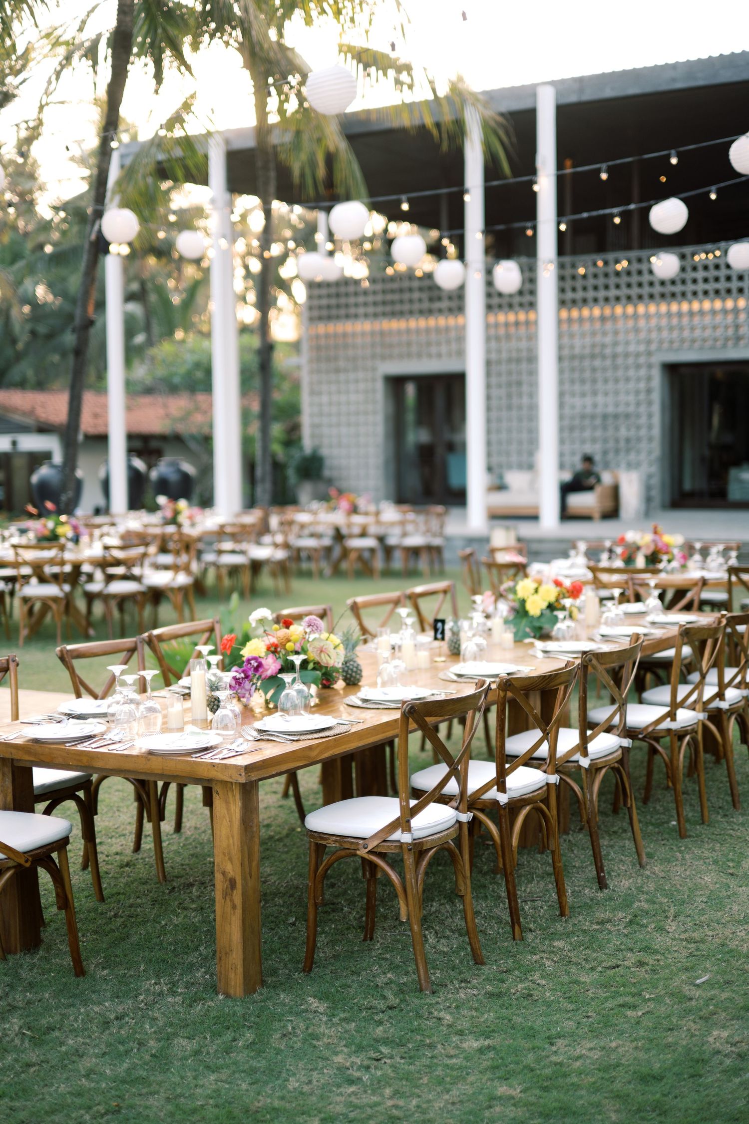 Outdoor wedding reception with wooden farm tables, colorful floral centerpieces, string lights, and white paper lanterns on green lawn.