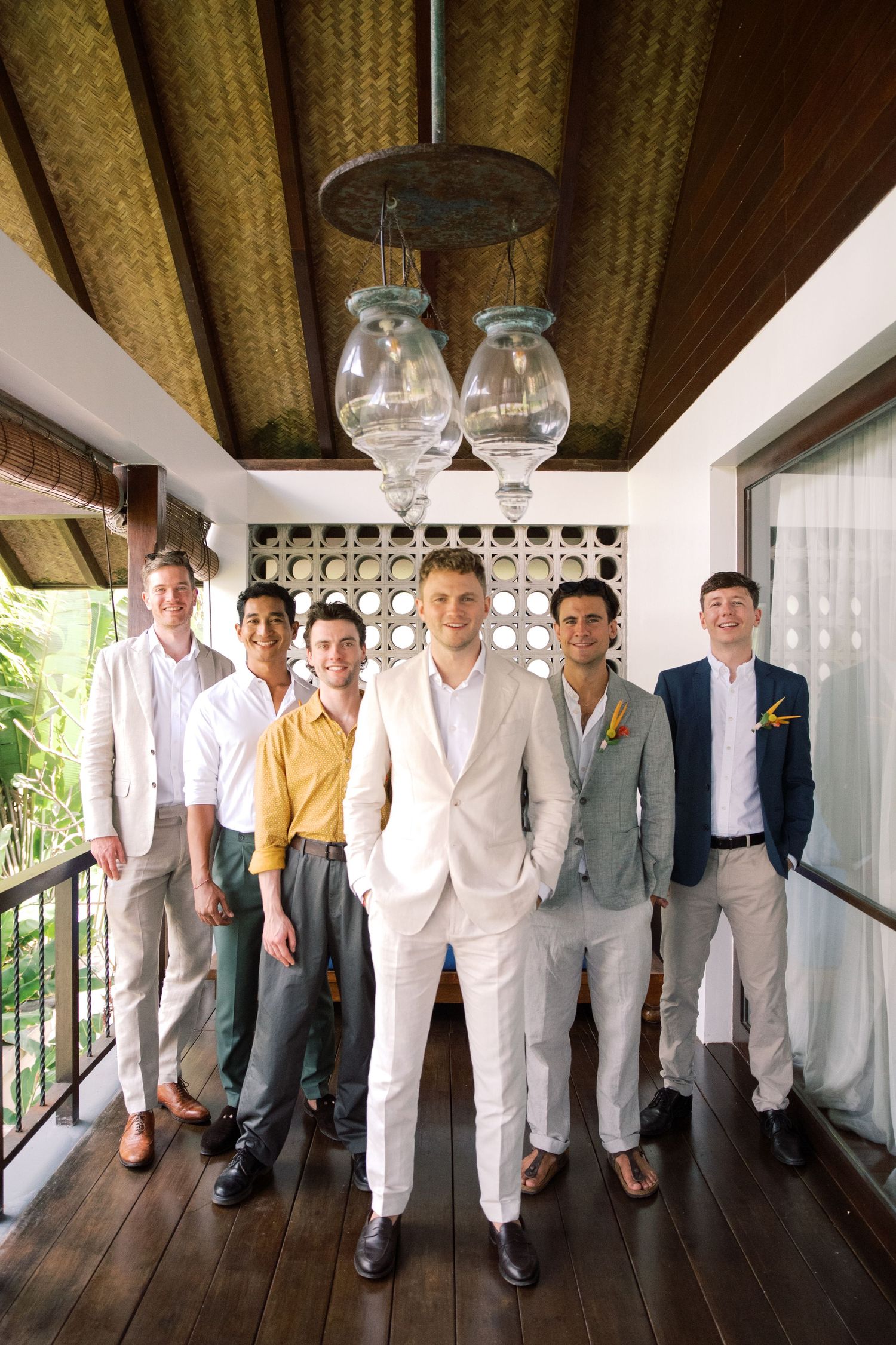 Groom in white suit poses with groomsmen in colorful attire at tropical wedding venue.