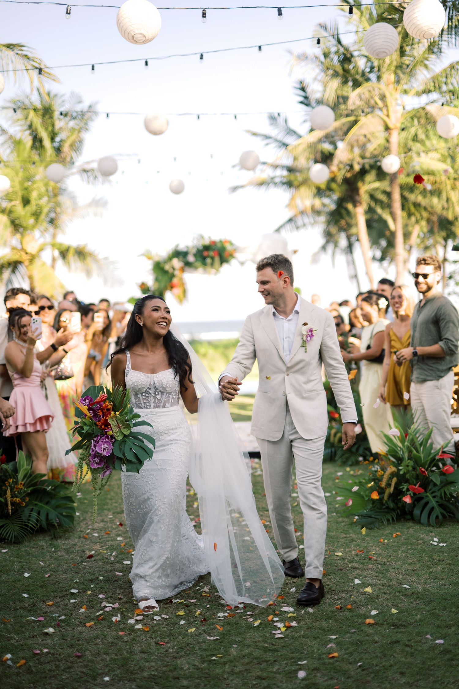 Newlyweds walk down aisle holding hands as guests celebrate under string lights and paper lanterns at outdoor wedding.