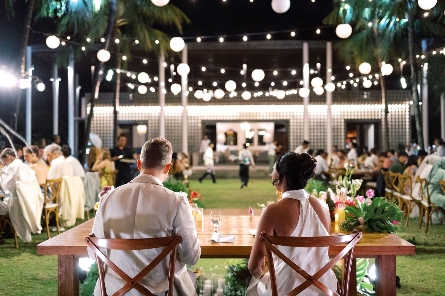Newlyweds sit at sweetheart table overlooking elegant outdoor tropical wedding reception with string lights and lanterns.