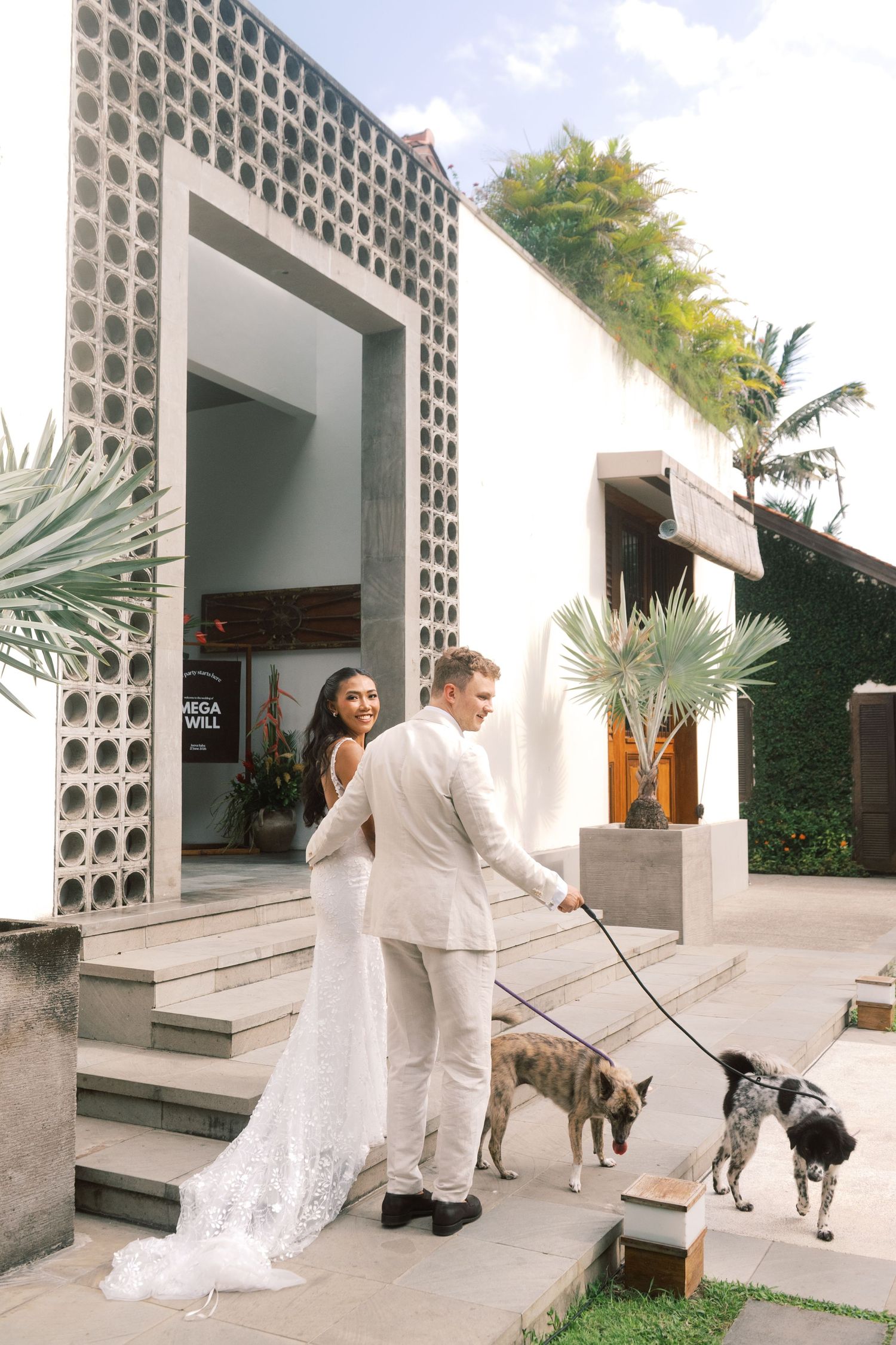 Bride and groom in white attire walking two dogs outside a modern tropical venue on their wedding day.