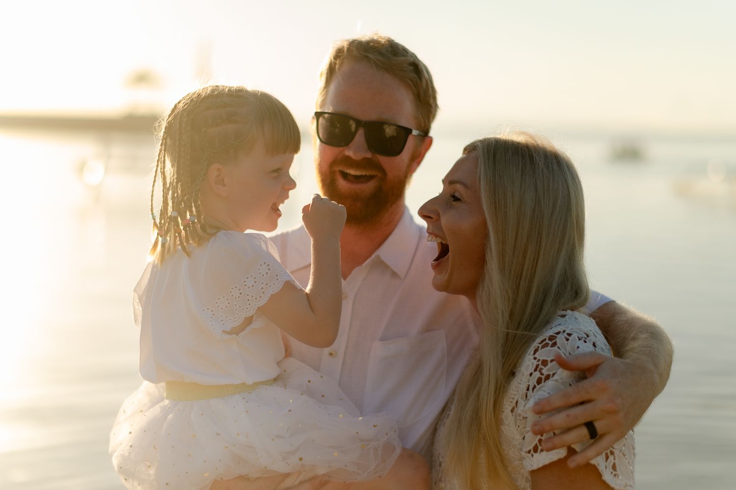 Smiling family with father holding young daughter as mother laughs on a bright sunlit beach.