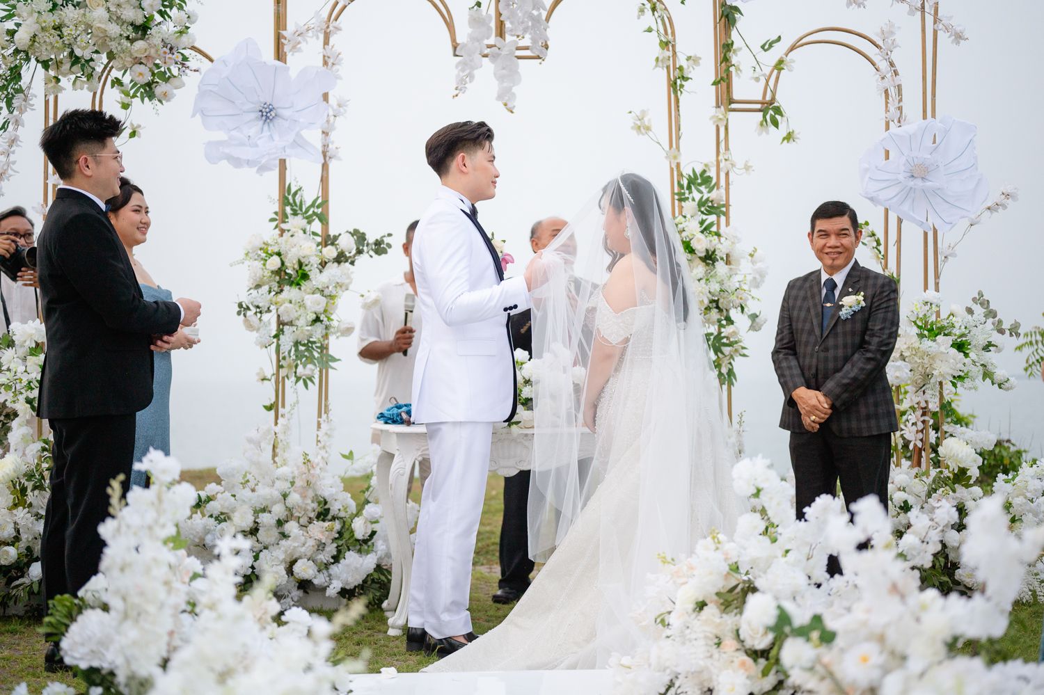 A groom in a white suit lifts his bride's veil during an elegant outdoor wedding ceremony adorned with white floral arrangements.