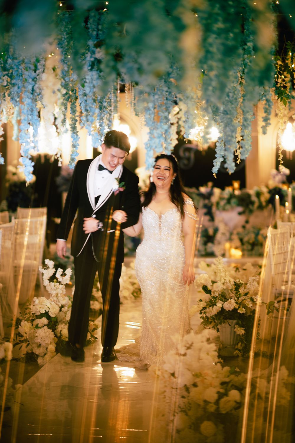 Joyful bride and groom walking down floral aisle with hanging blue wisteria and white flowers at elegant wedding reception.