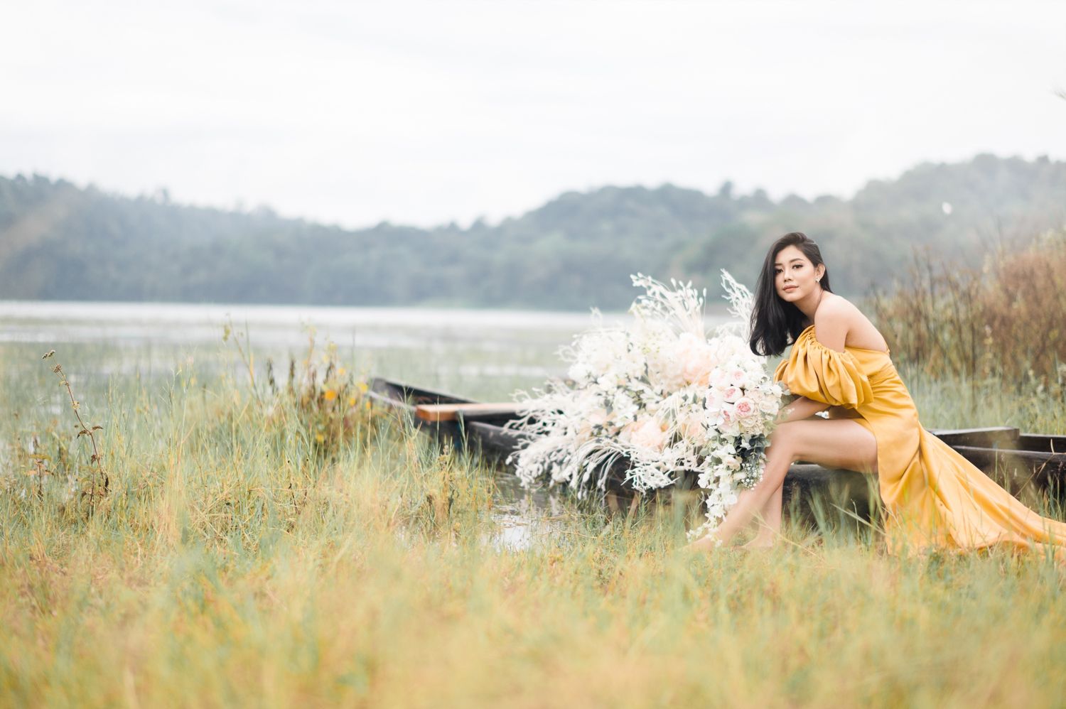 Woman in yellow off-shoulder gown sitting in boat surrounded by wildflowers near serene lake.