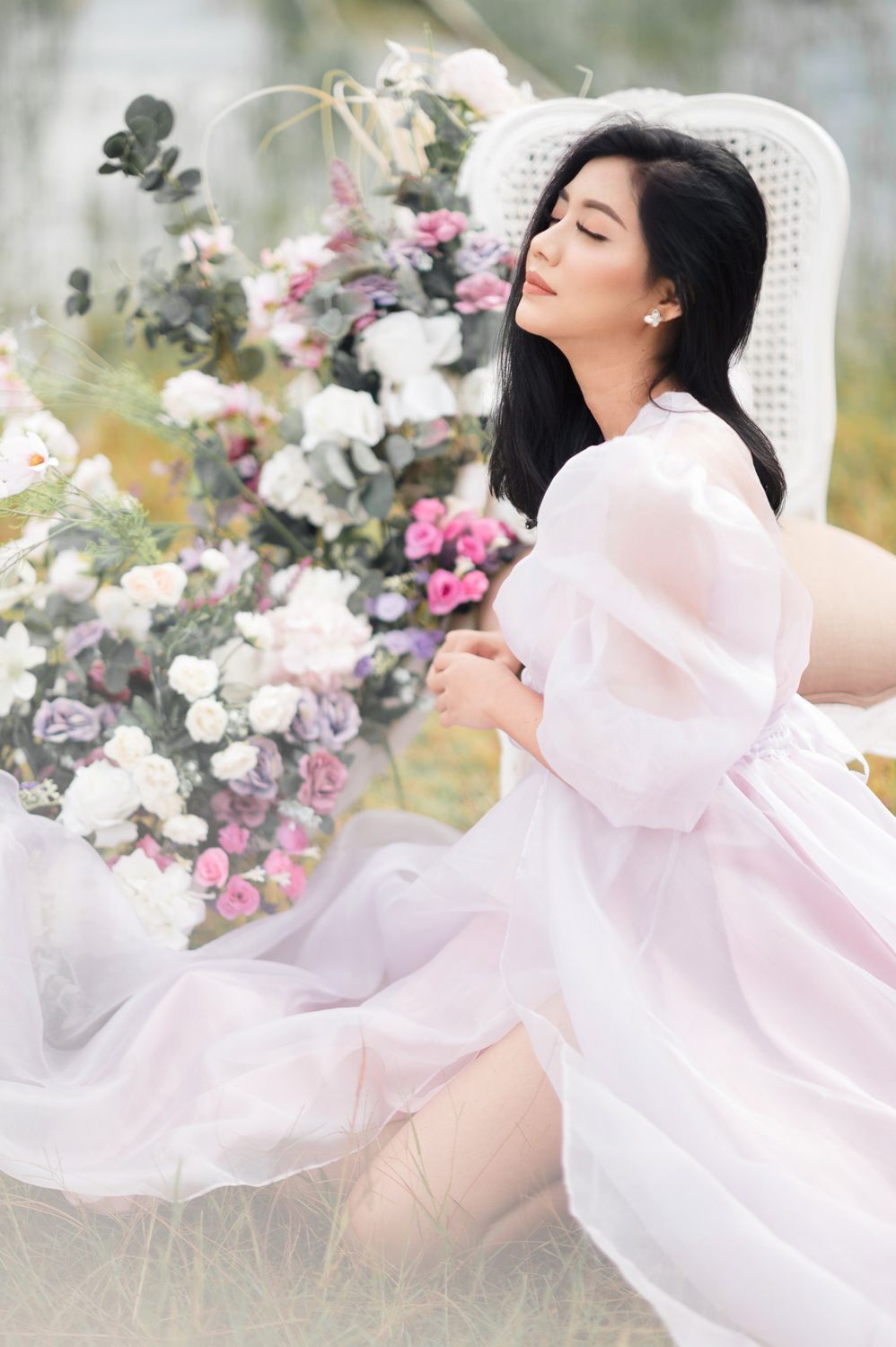 A woman in a flowing pink organza dress sits gracefully outdoors among lush floral arrangements of white and purple blooms.