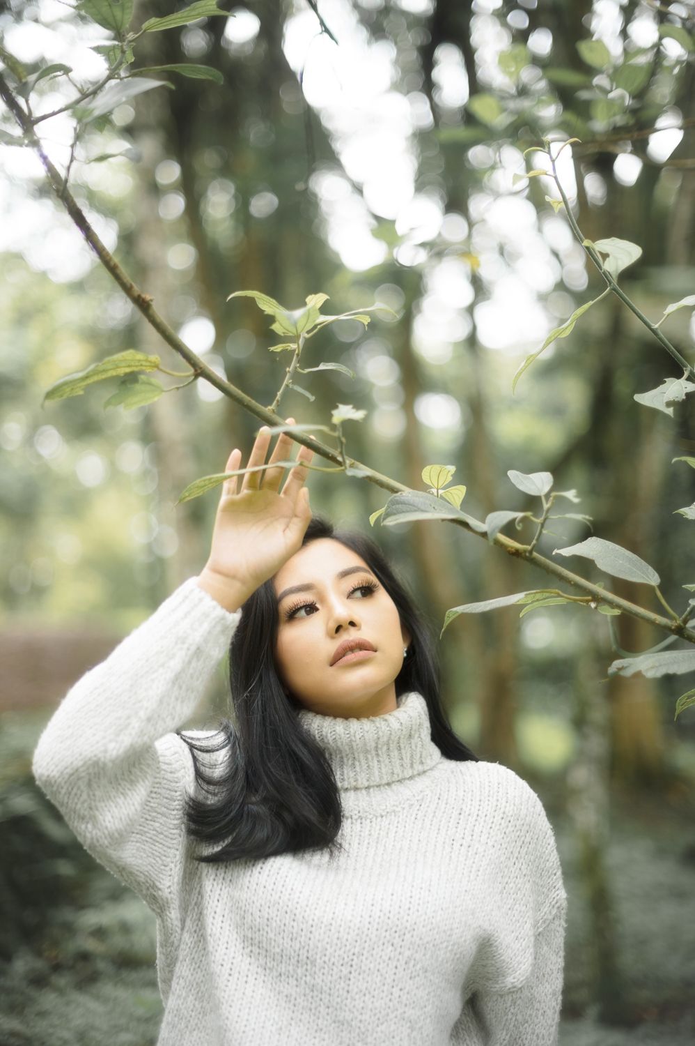 A young Asian woman in a white turtleneck sweater poses gracefully among green forest branches.