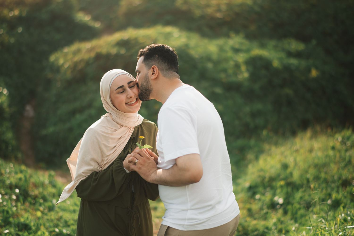 A Muslim couple sharing a tender moment outdoors, woman in hijab smiling as man leans in close, green nature background.