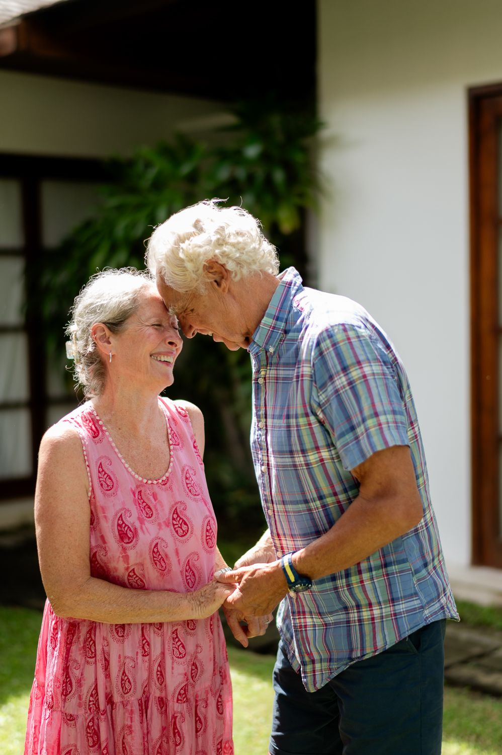 Elderly couple holding hands and touching foreheads while smiling outdoors near a white building.