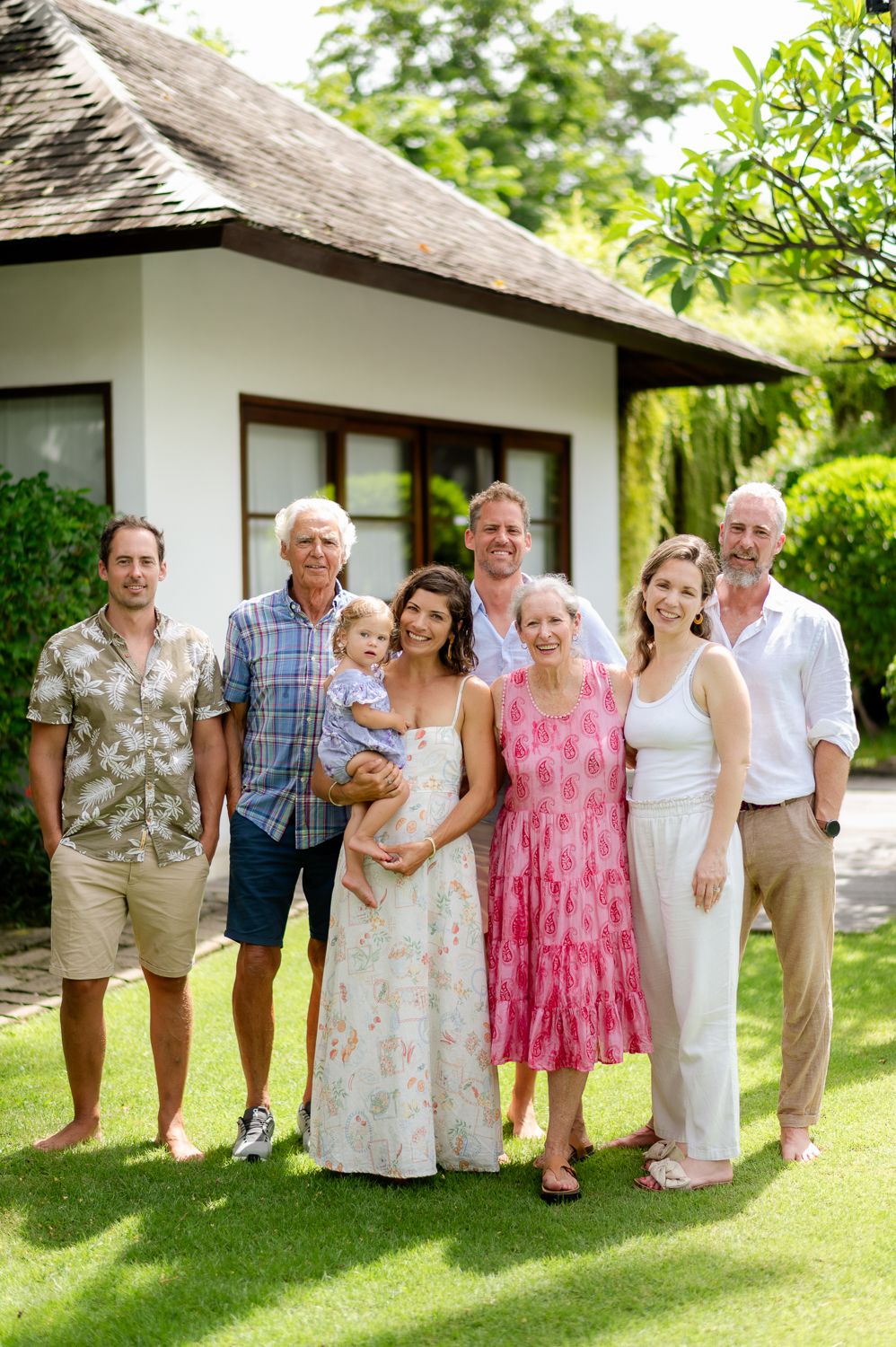 Multi-generational family smiling together outdoors on green lawn in front of tropical villa.