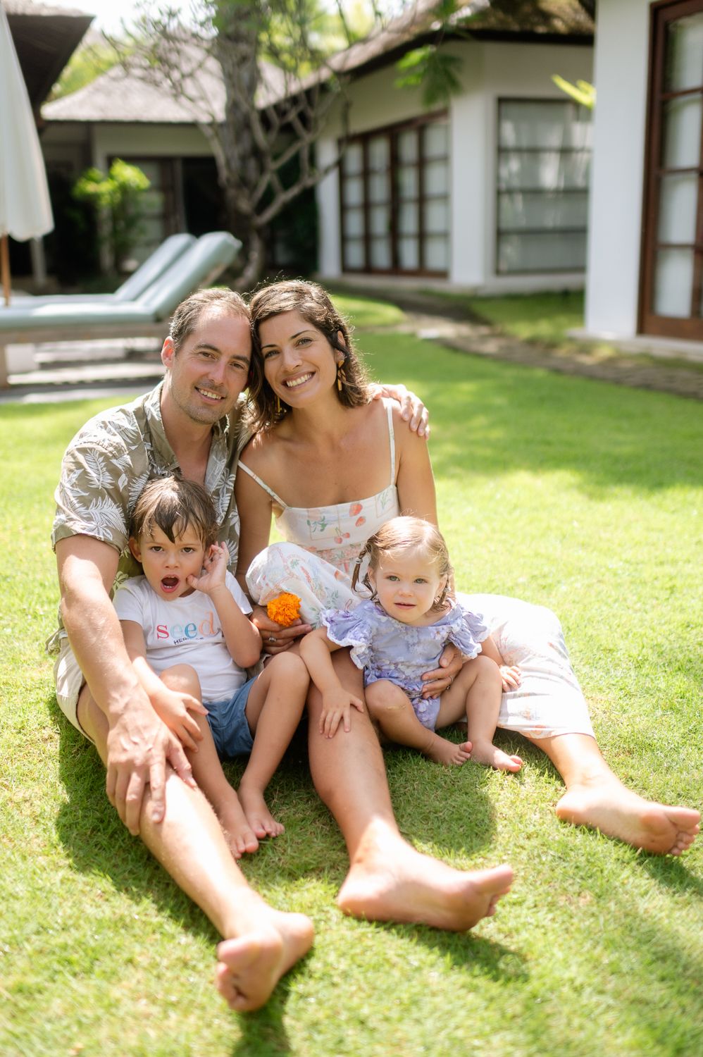 Happy family of four sitting on green grass in front of a tropical villa, smiling parents with two young children.