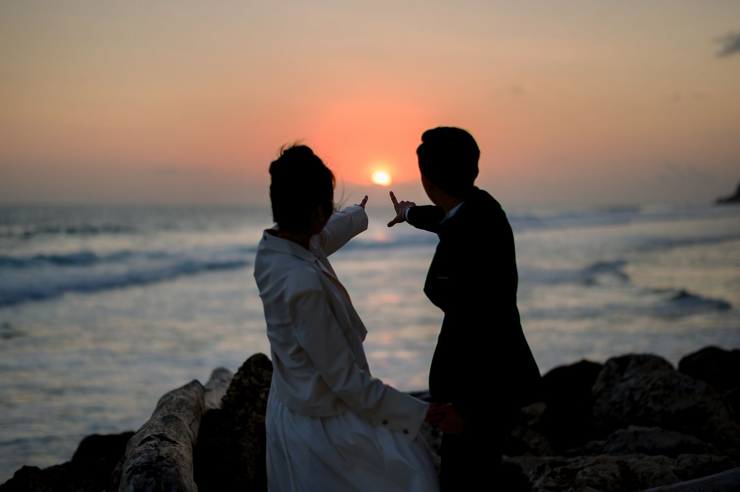 Silhouette of a couple in wedding attire posing at sunset on rocky shore with ocean waves behind them.