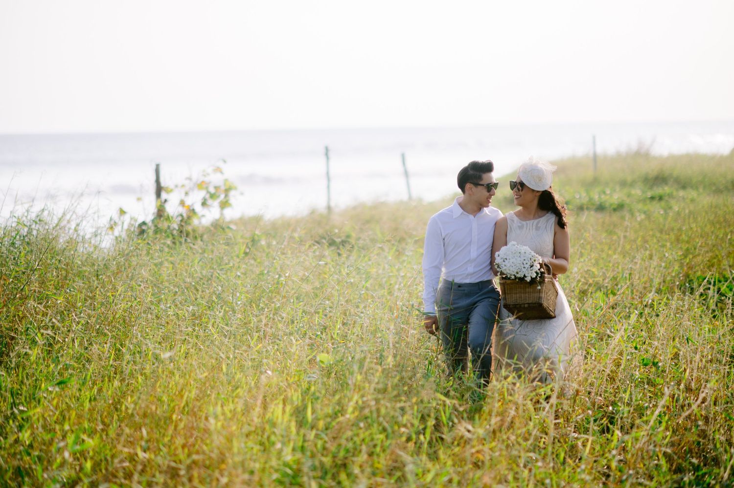 Couple walking through tall grass field near ocean shore, woman in white dress and hat holding bouquet.