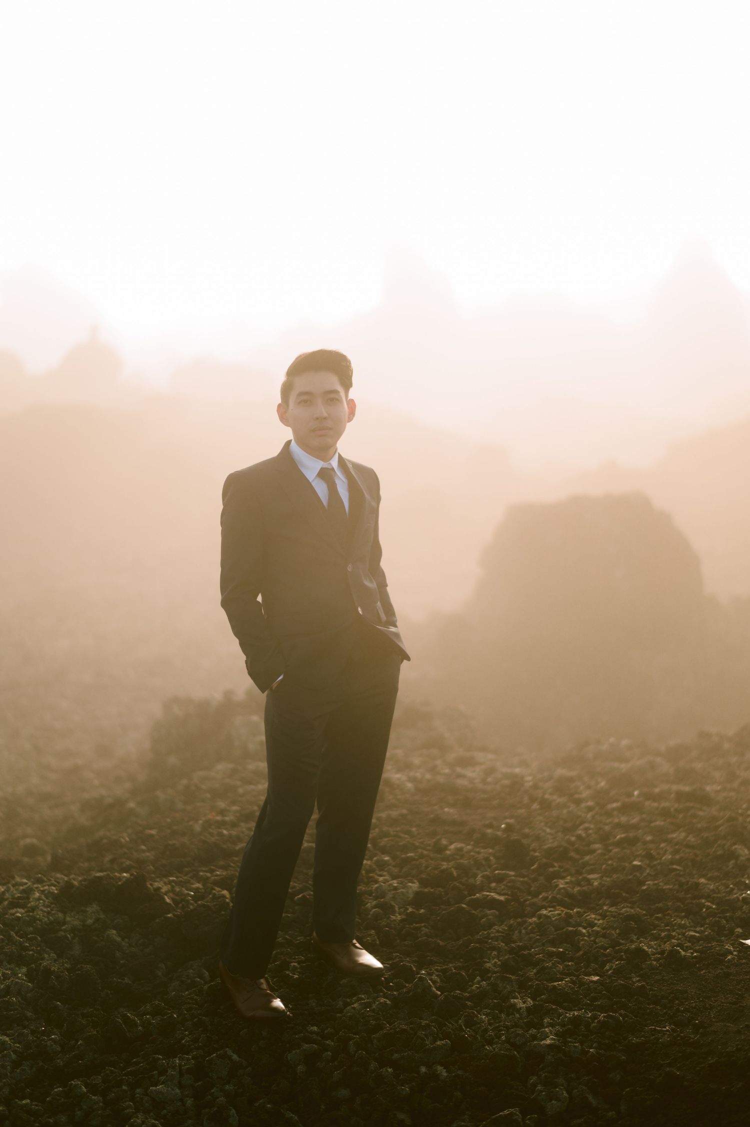 Young man in dark suit and tie standing in misty foggy field at golden hour, professional portrait photography.