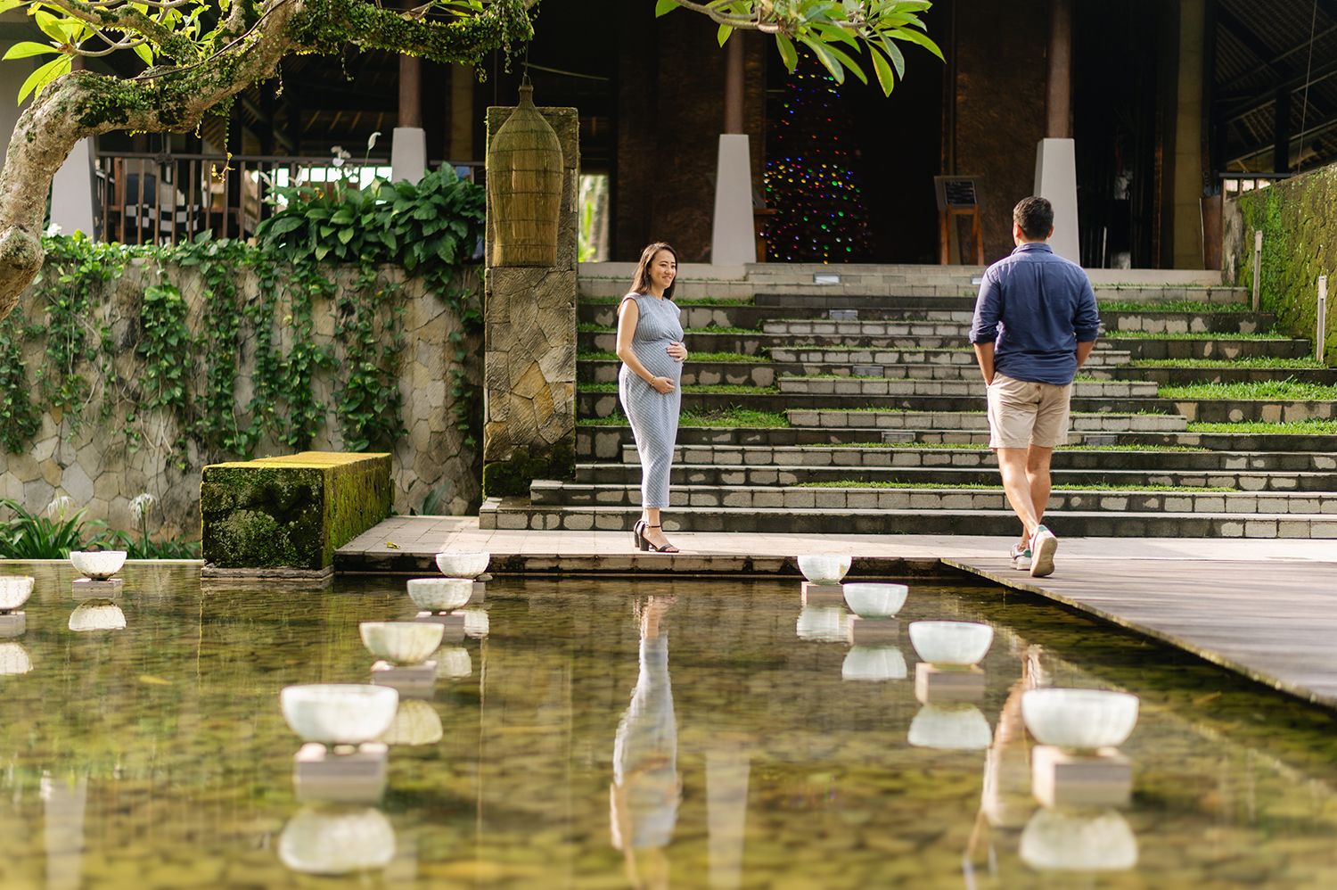 Couple standing by a serene reflective pool with stepping stones in a lush tropical garden setting Maya Ubud Resort and Spa in Bali.