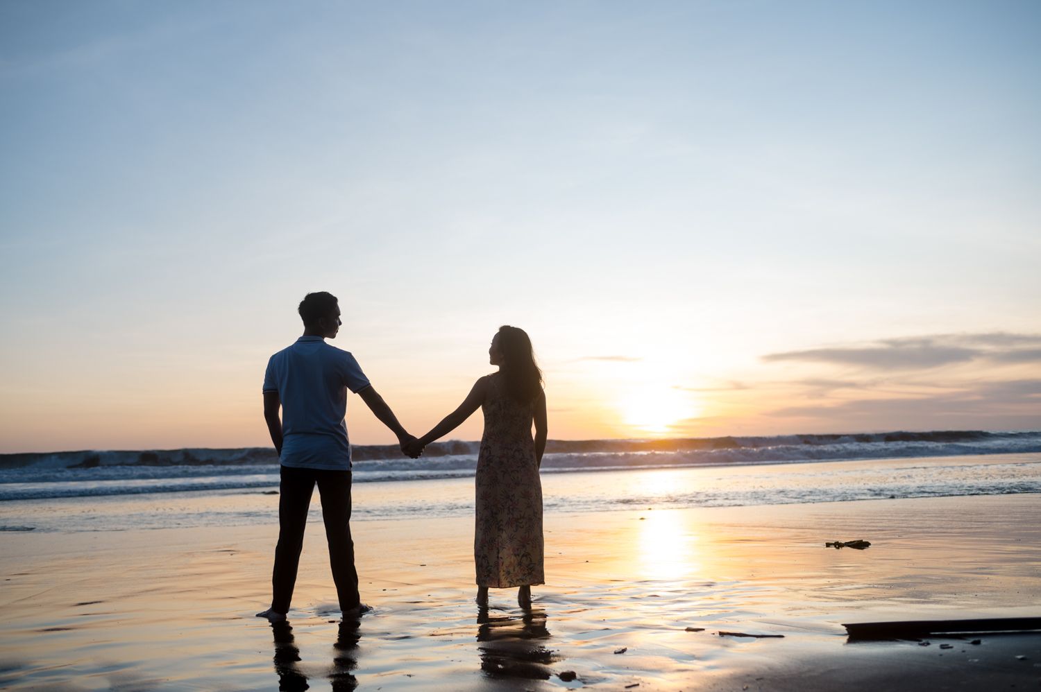 Couple holding hands silhouetted against a stunning ocean sunset on a wet reflective beach.