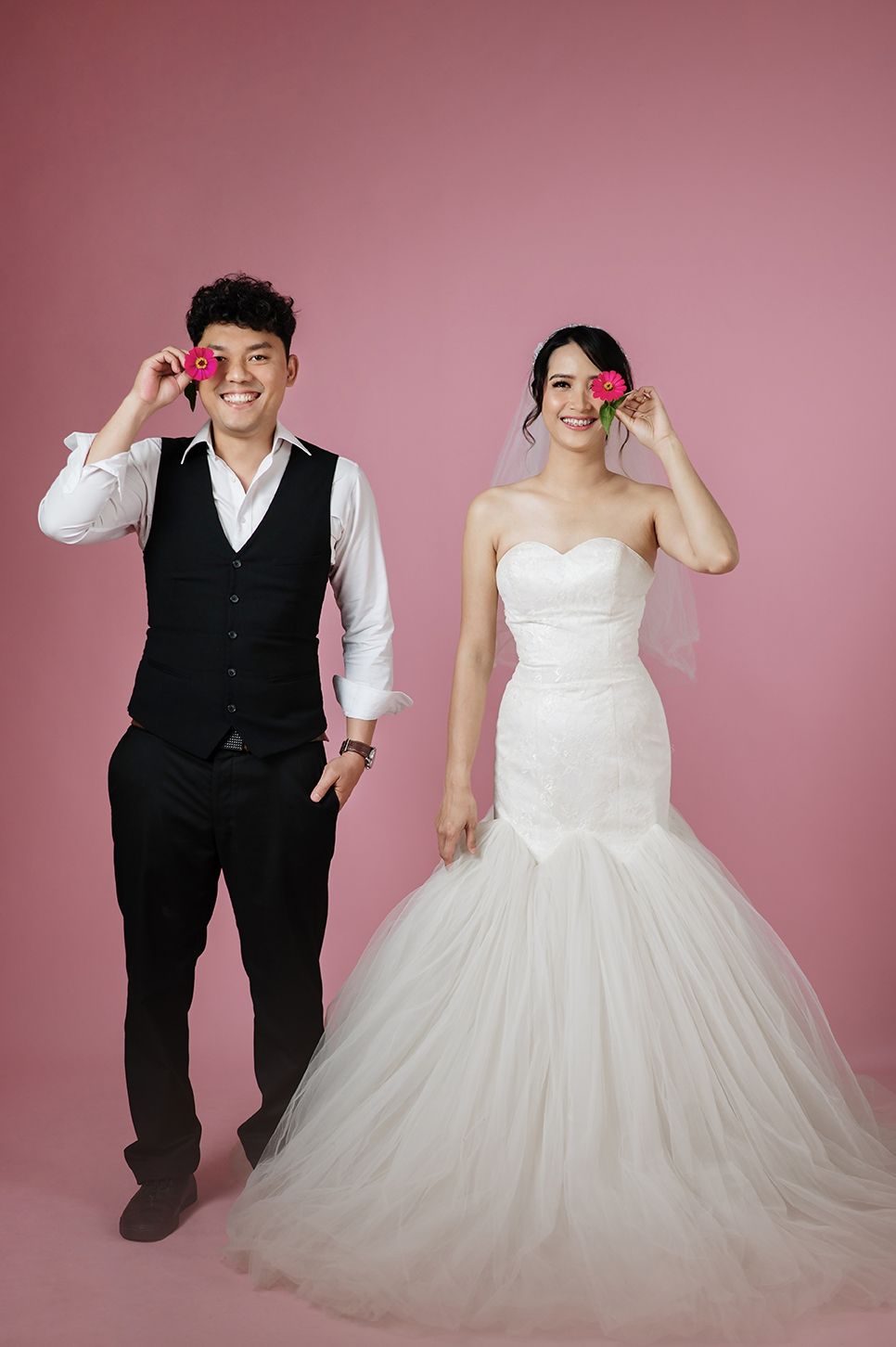 A smiling couple in wedding attire pose playfully with pink roses against a pink background.