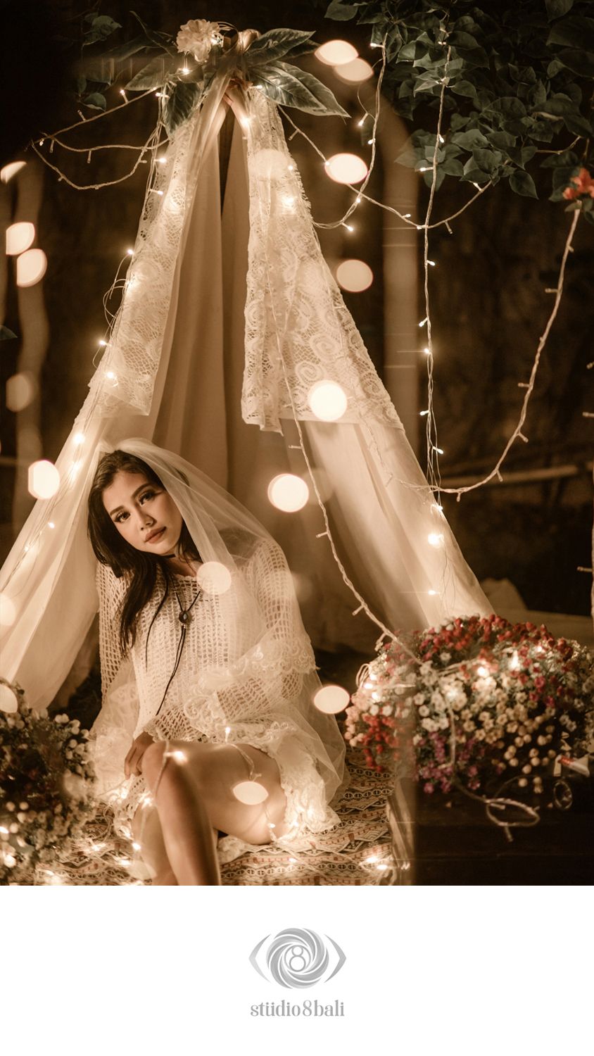 Bride in lace gown sits under glowing fairy light canopy with floral arrangements in romantic Bali studio setting.