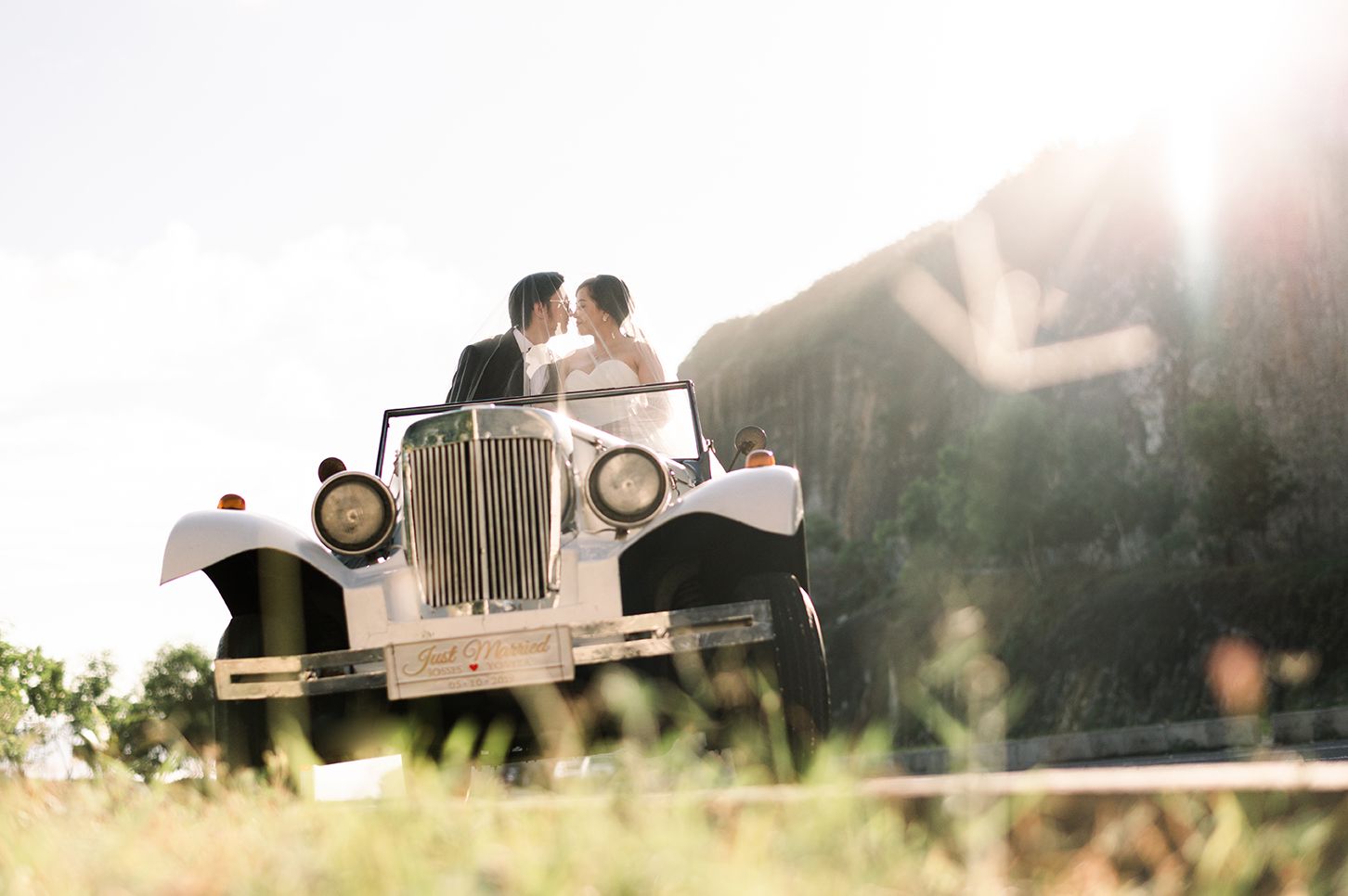 Bride and groom kissing behind a vintage white classic car on a sunny wedding day outdoors.