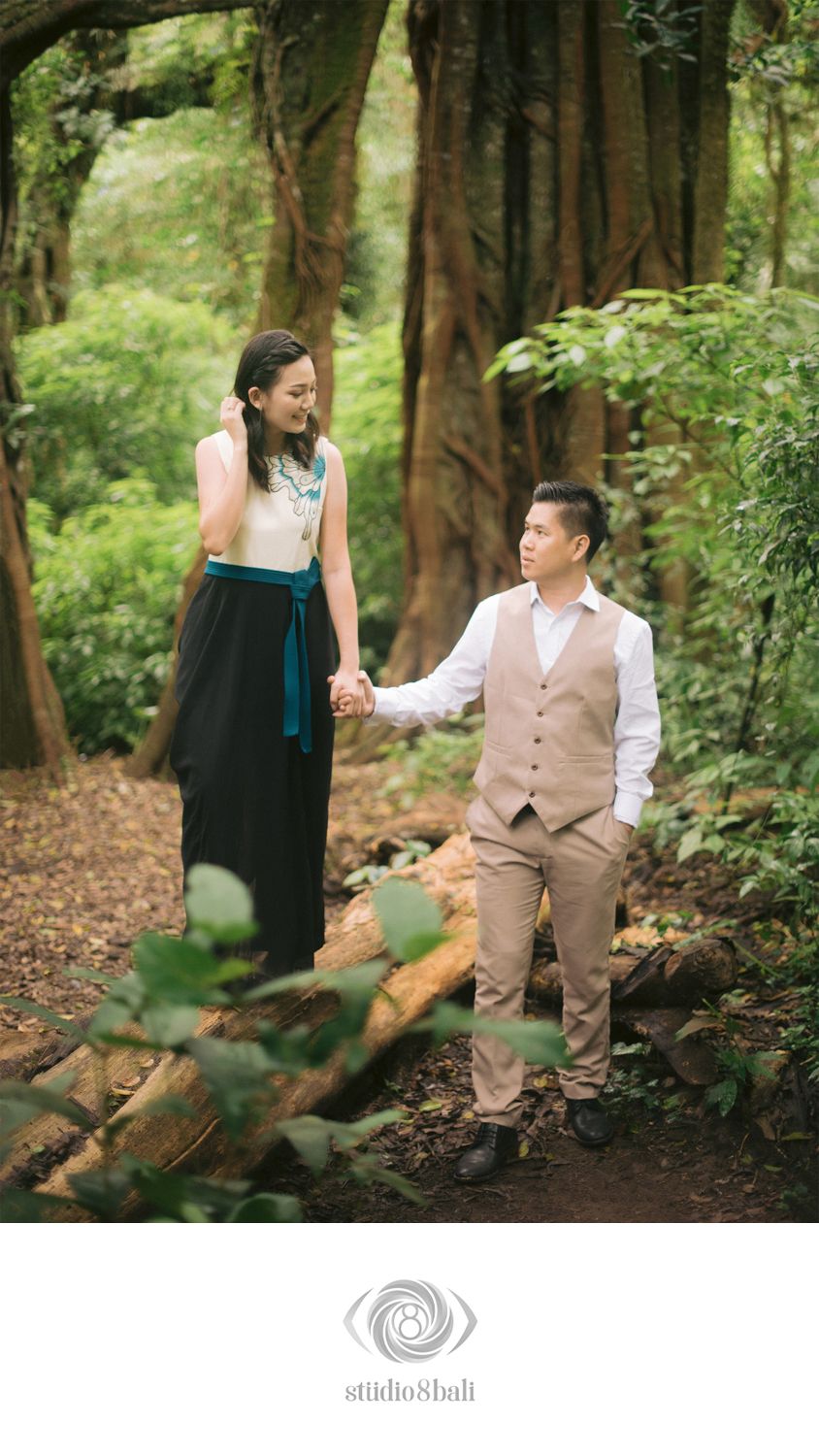 Couple holding hands in a lush green forest, woman in black skirt and man in tan vest, romantic outdoor portrait.