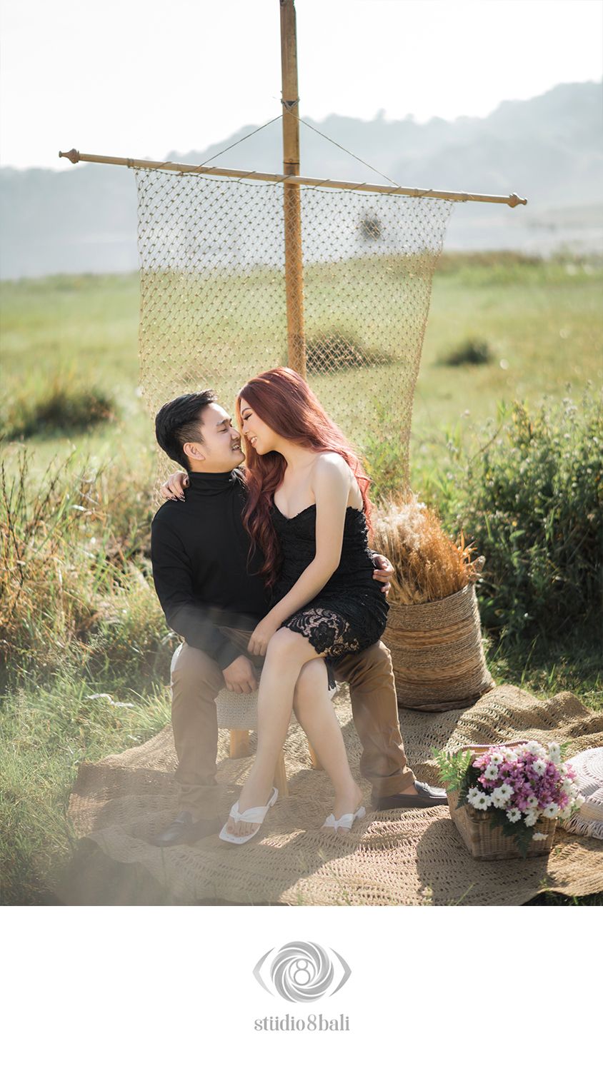 Couple sitting together on a rustic mat in a scenic field, surrounded by flowers and a decorative bamboo structure.