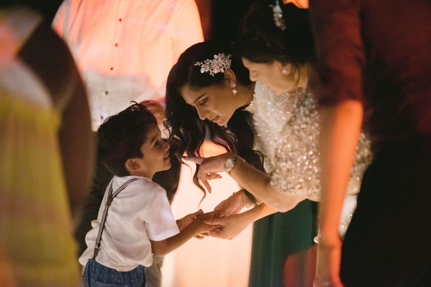 A bride in a sequined gown and hair accessory interacts warmly with a young boy in suspenders at a wedding reception.