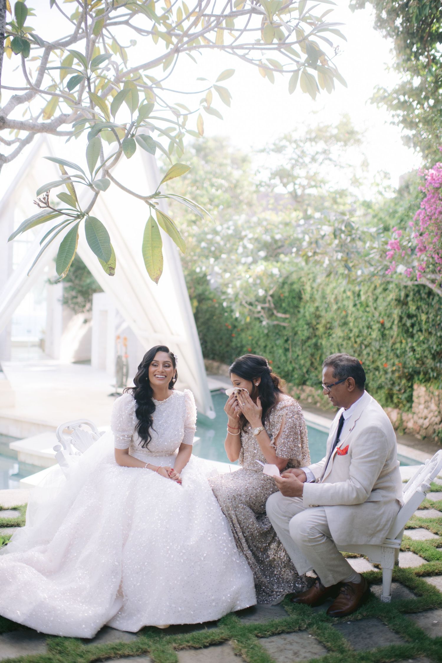 Bride in white gown sits with family by pool in lush garden setting during an outdoor wedding celebration.