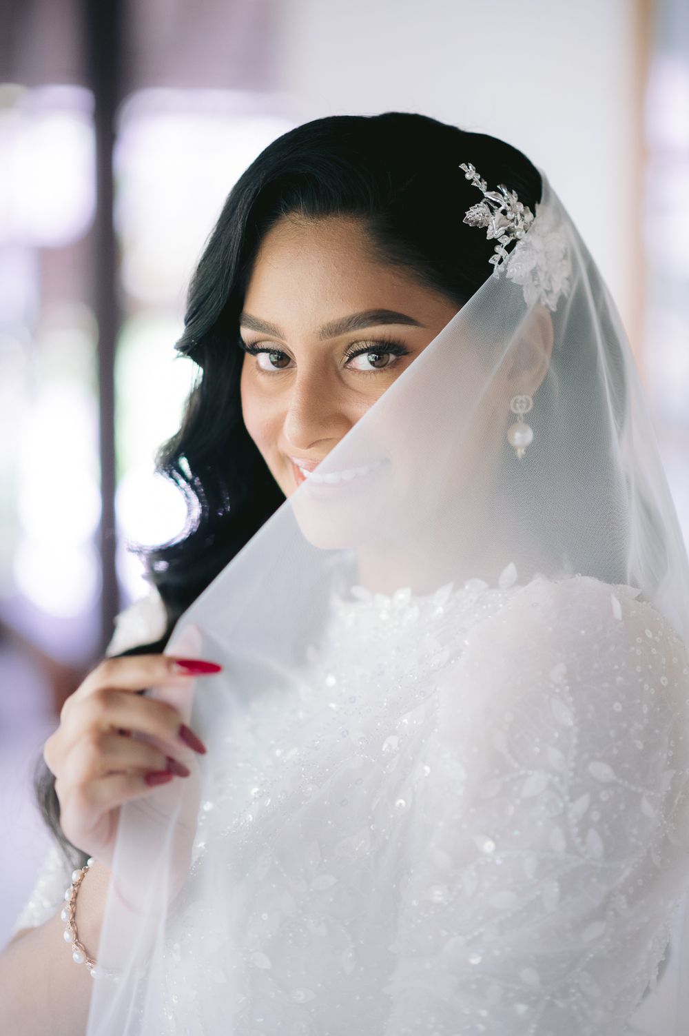 A smiling South Asian bride in a white lace gown holds her embroidered veil up to her face, wearing pearl earrings.