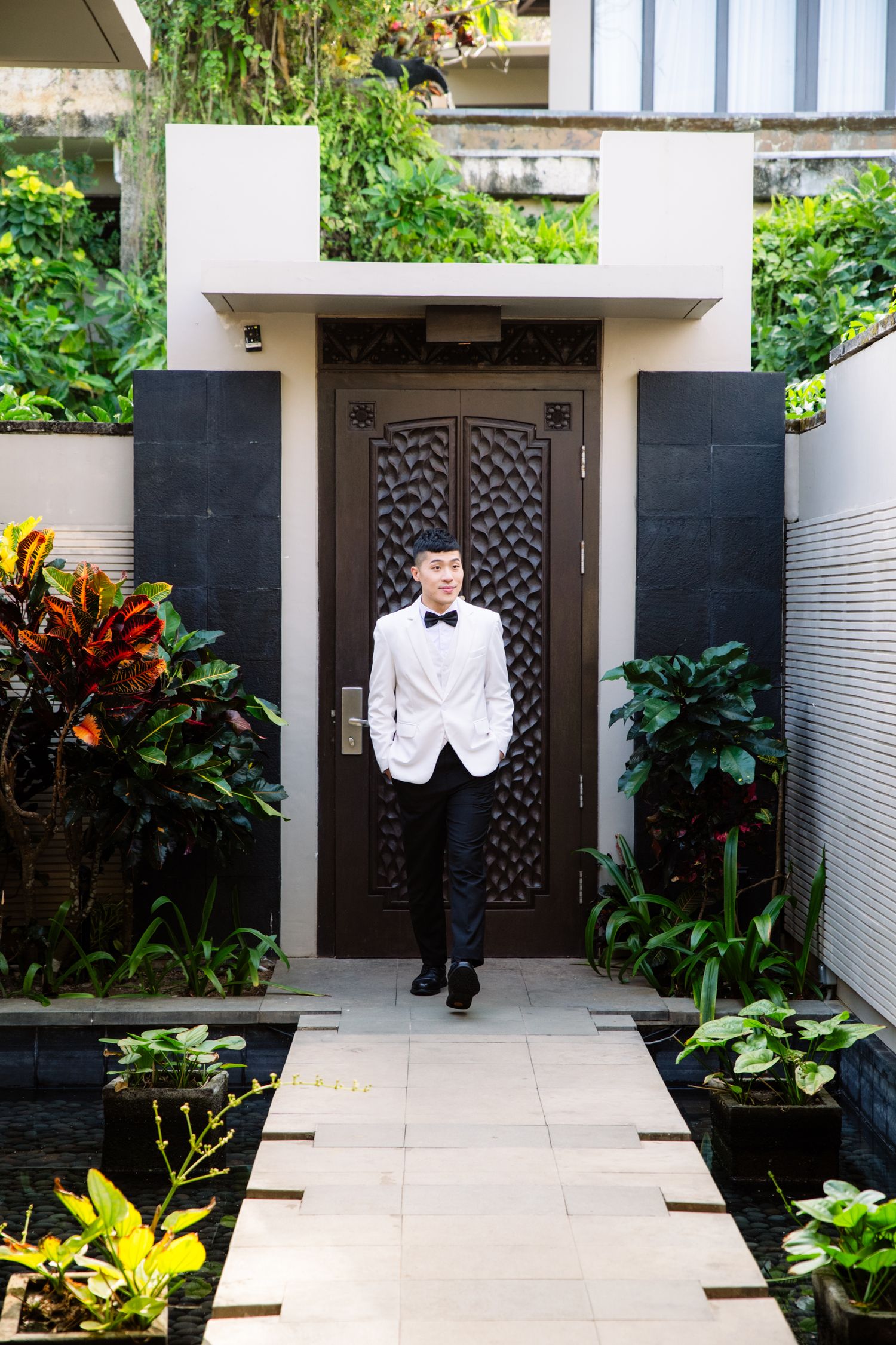 Man in a white tuxedo jacket and black bow tie walks along a stone path toward an ornate wooden door, surrounded by lush tropical plants.