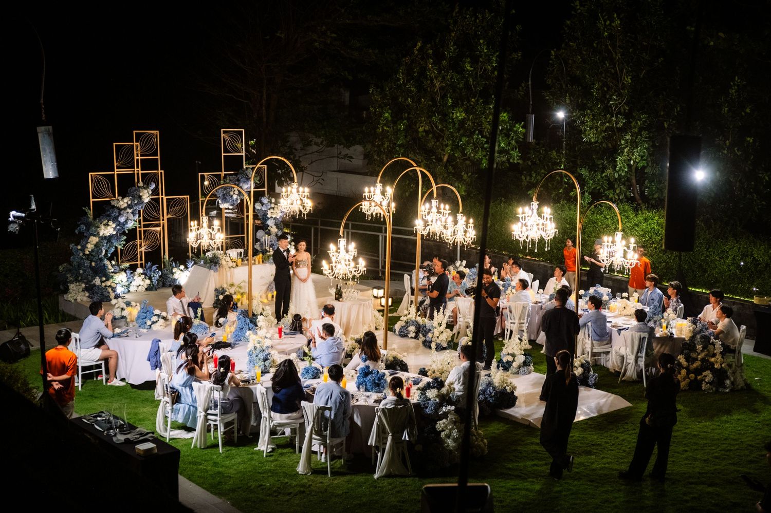 Elegant outdoor evening wedding reception on a lawn with guests seated at round tables beneath glowing gold chandeliers and floral decor.