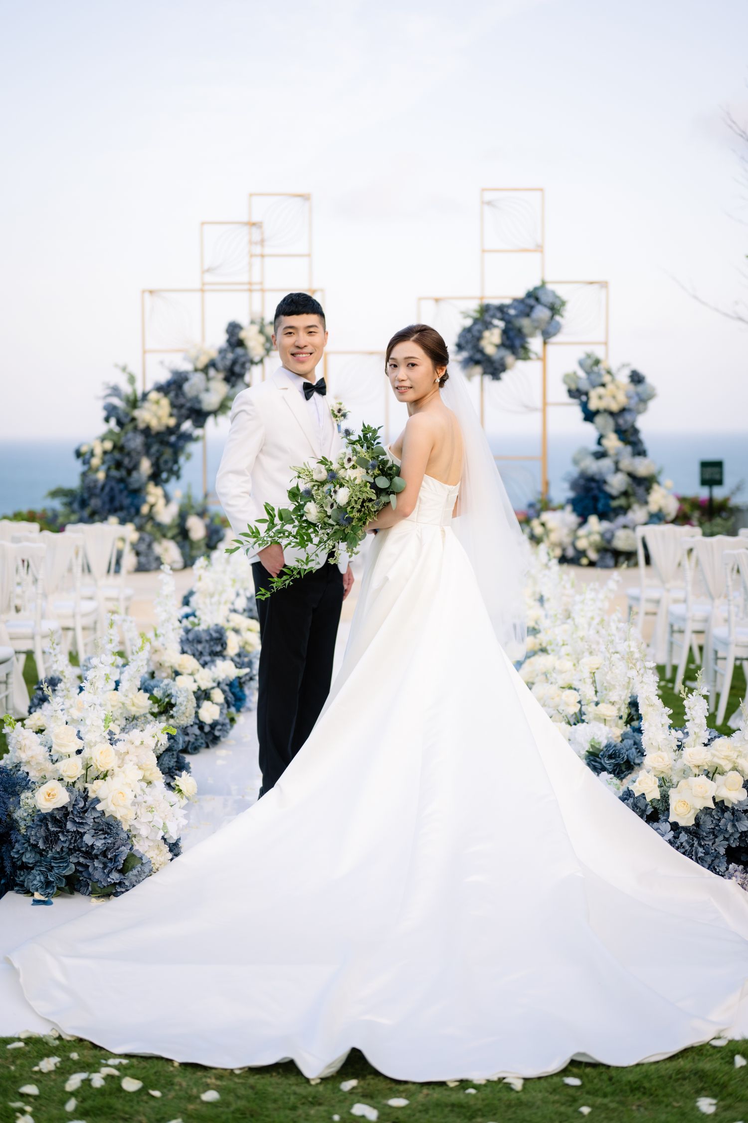 Bride in a flowing white gown and groom in a white tuxedo pose with a bouquet at an oceanside wedding adorned with blue and white flowers.