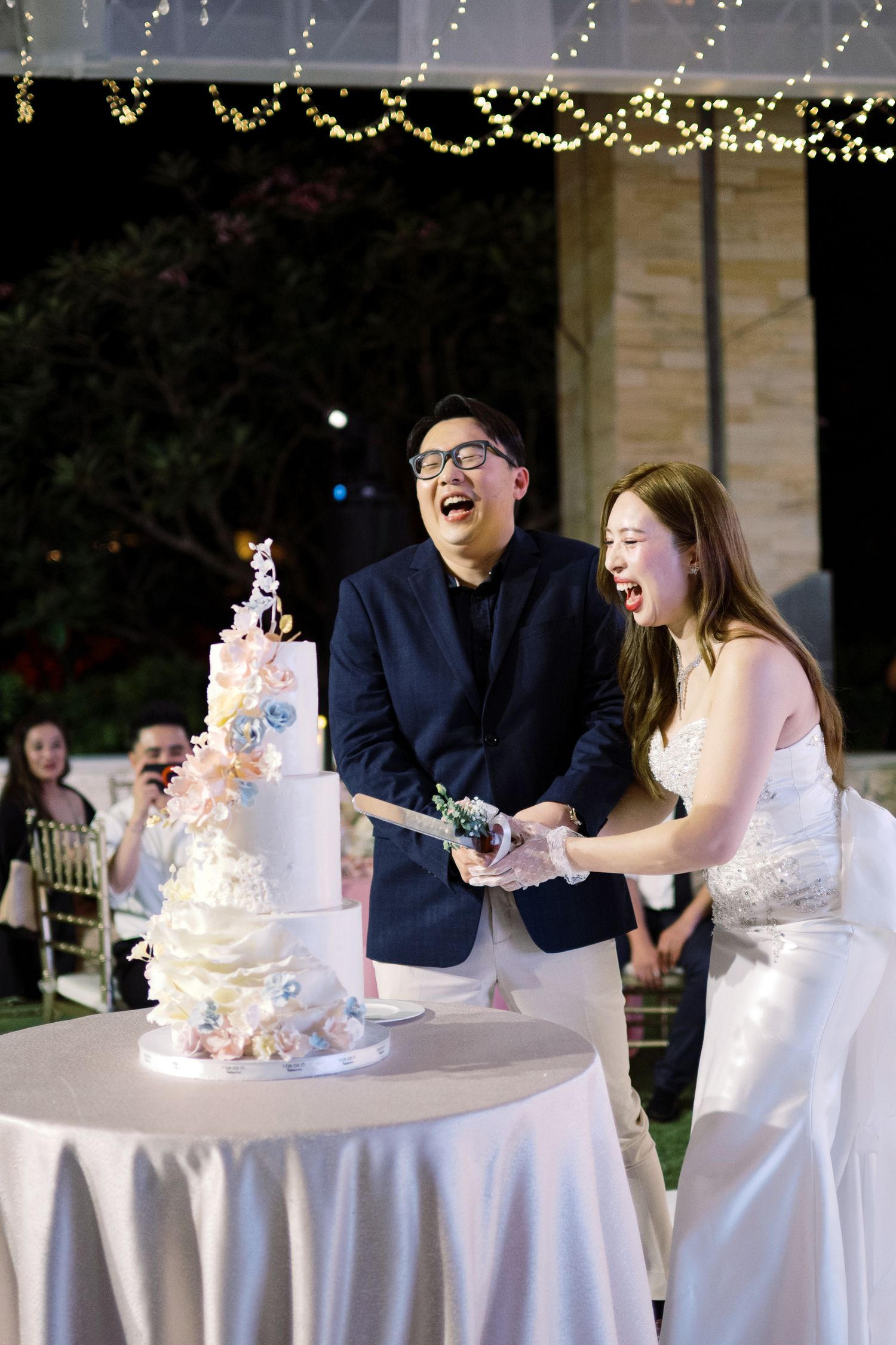 Joyful couple laughing while cutting a white floral wedding cake at an outdoor evening reception.