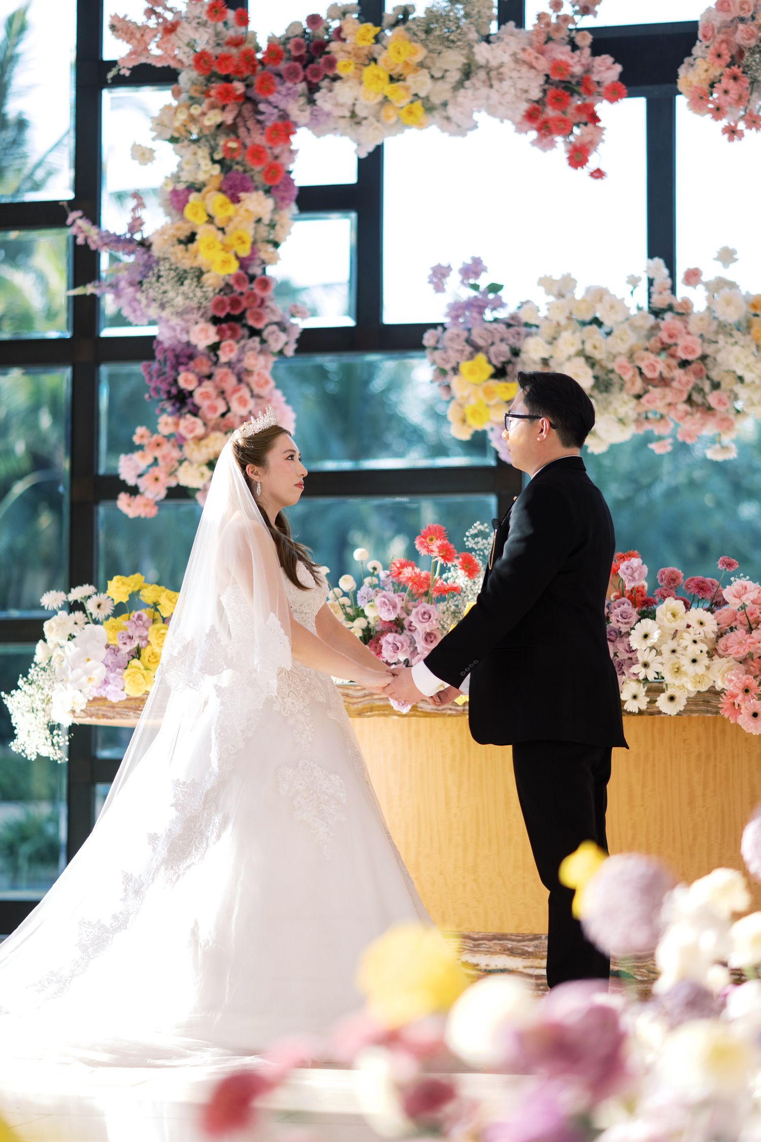 Bride and groom exchange vows at a colorful floral wedding ceremony altar with vibrant pink, yellow, and white flowers.