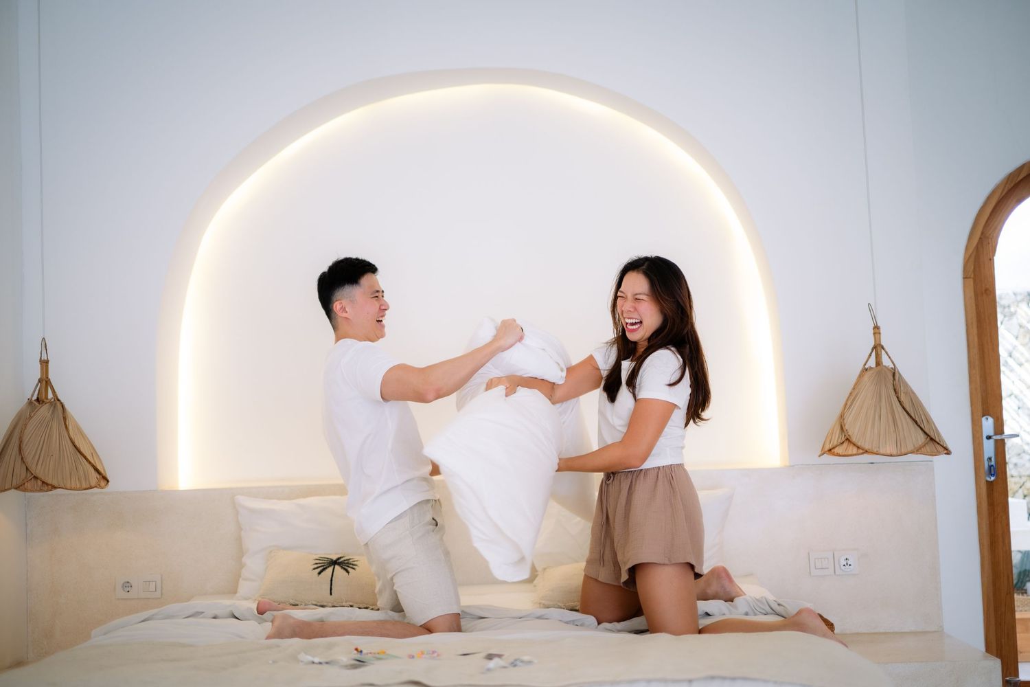 Young couple playfully having a pillow fight on a white bed in a modern bedroom with arch lighting.