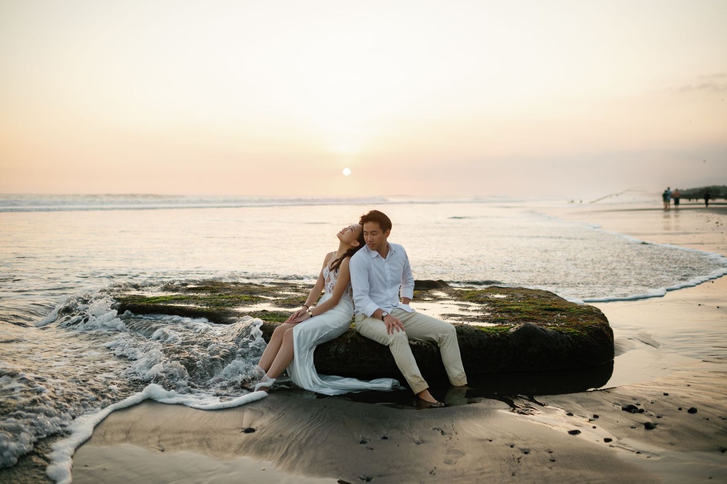 Couple sitting on a rock at the beach during a romantic sunset, waves gently washing around them.