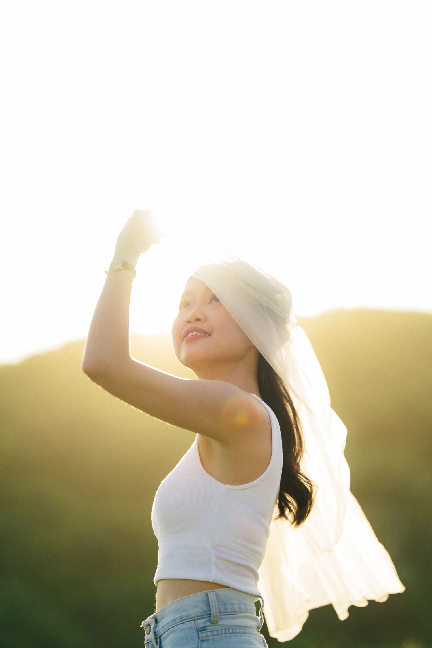 Young Asian woman in white crop top and hat poses outdoors at golden sunset with arms raised, mountains in background.