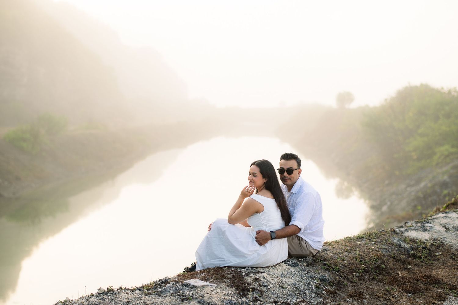Couple in white sitting on rocky cliff overlooking misty river, romantic outdoor portrait.