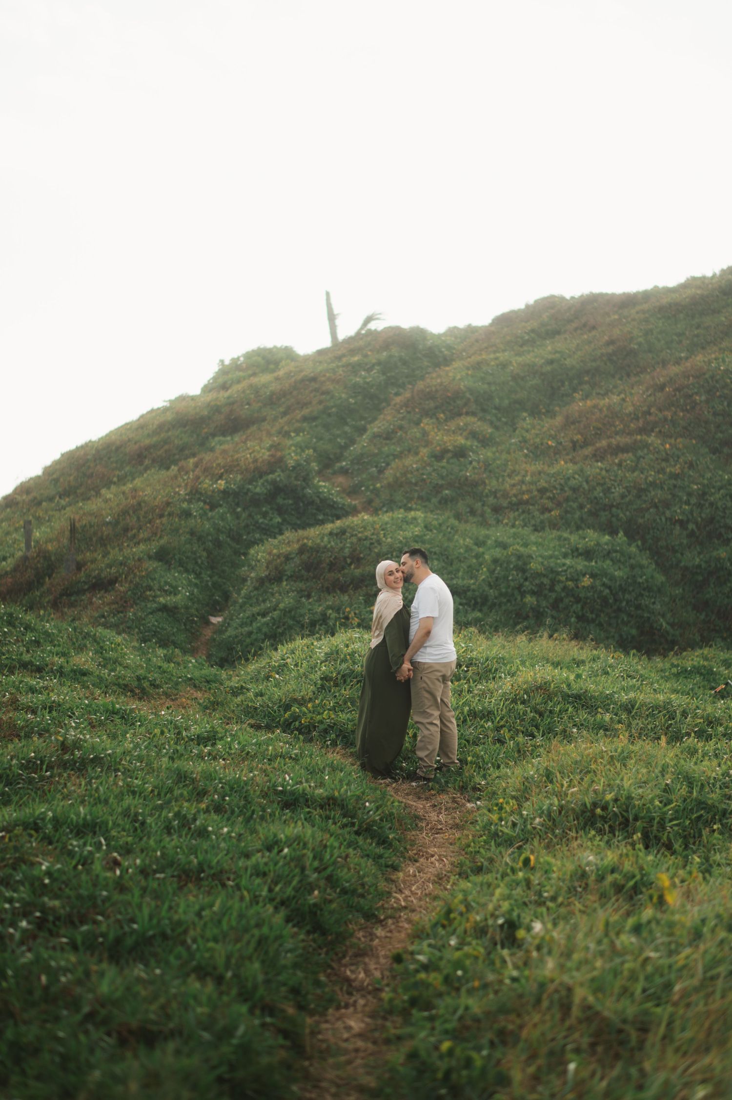 A couple embraces on a lush green hillside trail, the woman in a dark dress and man in white shirt and khaki pants.
