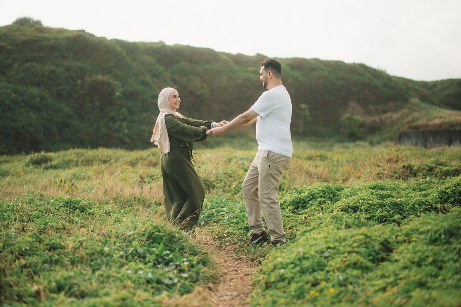 A couple in traditional attire holds hands on a green hillside path, sharing a romantic outdoor moment.