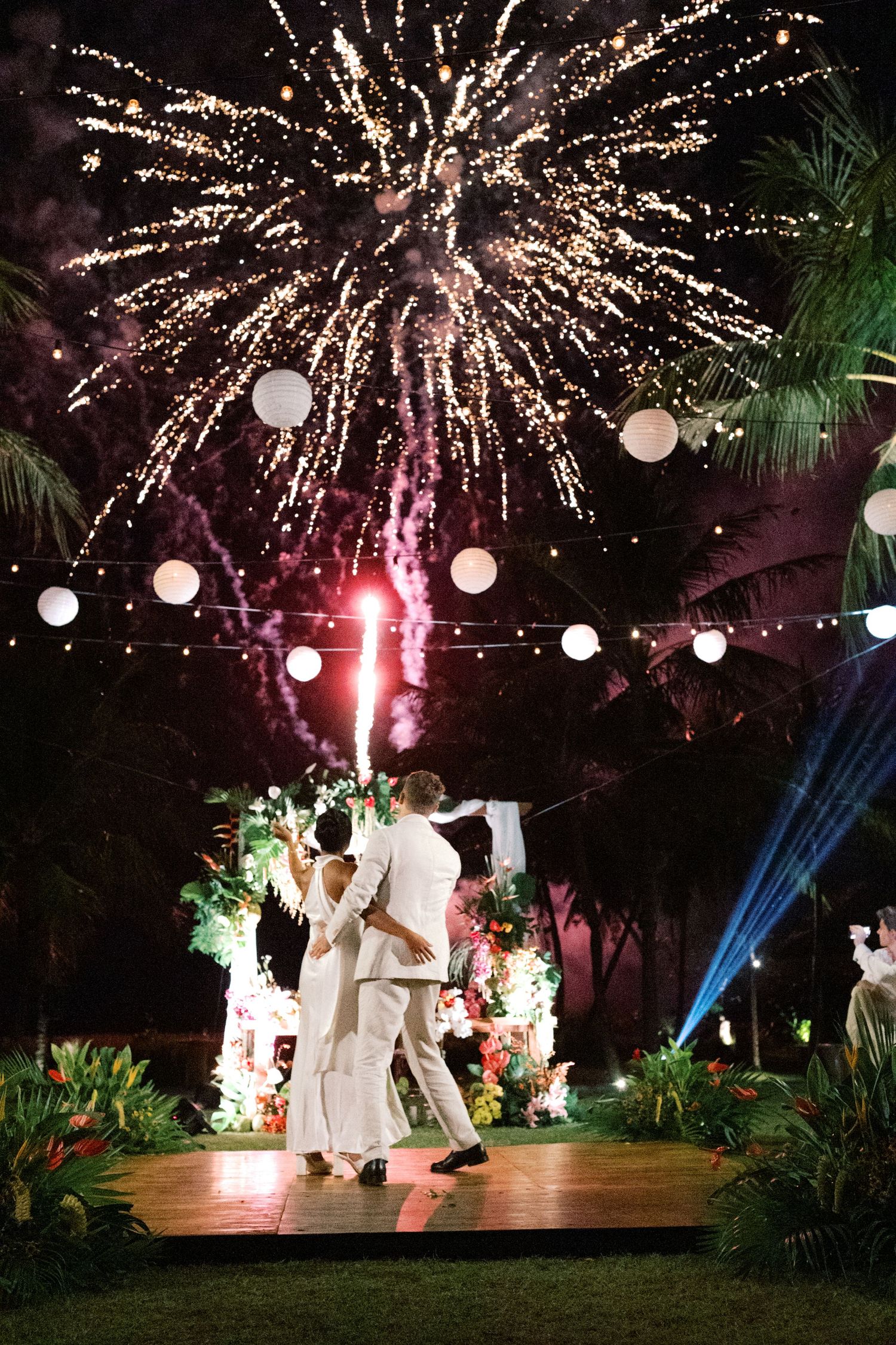 Bride and groom share first dance on outdoor wooden floor with stunning fireworks exploding overhead at night.