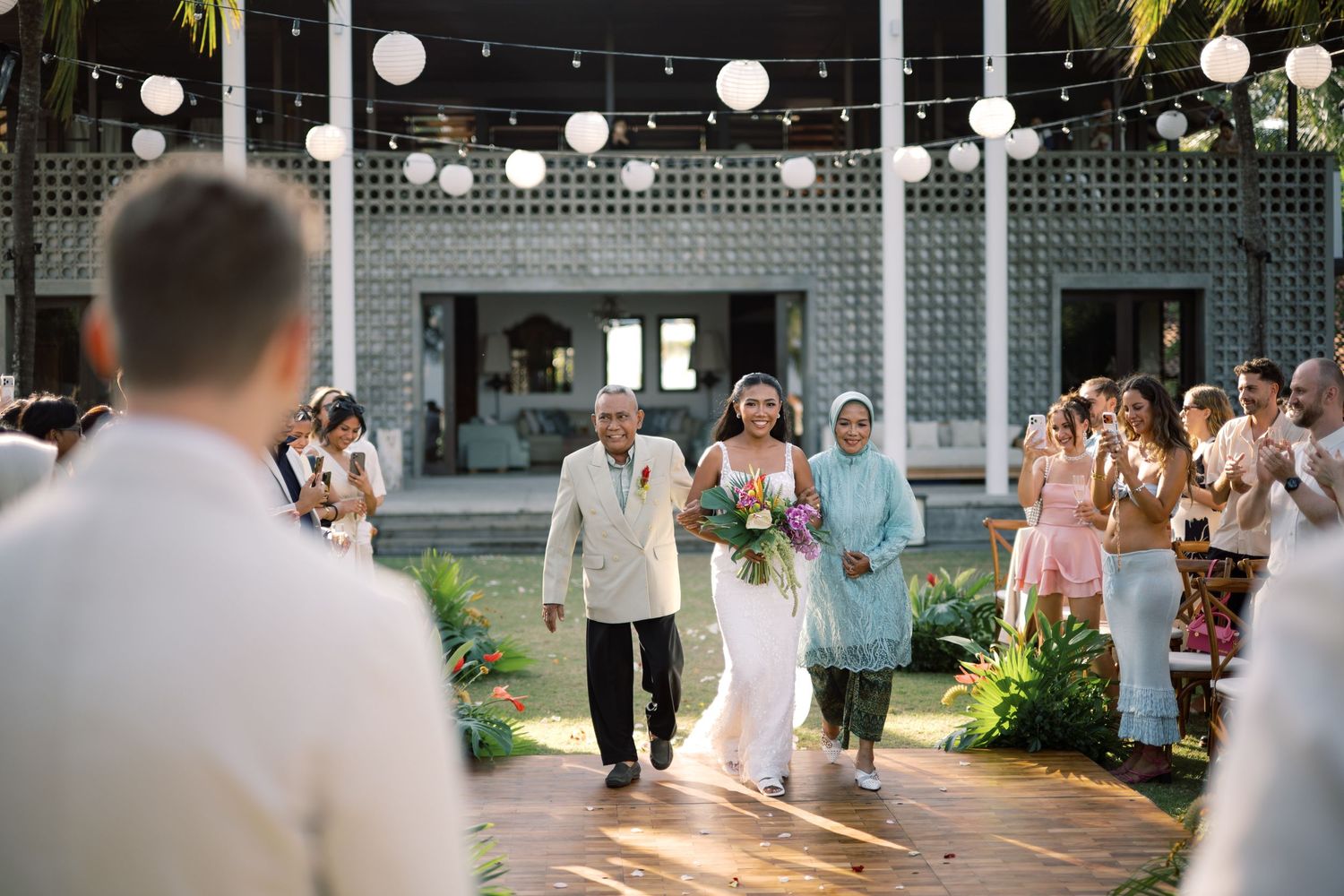 Bride walks down outdoor aisle with elderly man as guests celebrate at a tropical wedding.