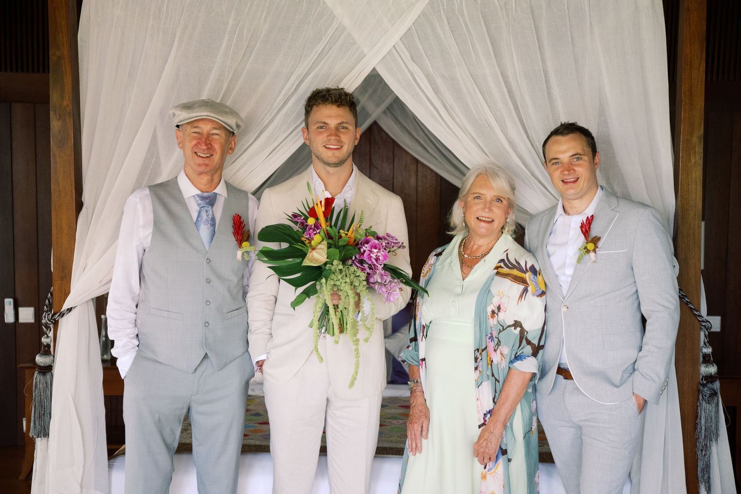 Groom with family posing in light suits and floral dress before tropical canopy backdrop at wedding.