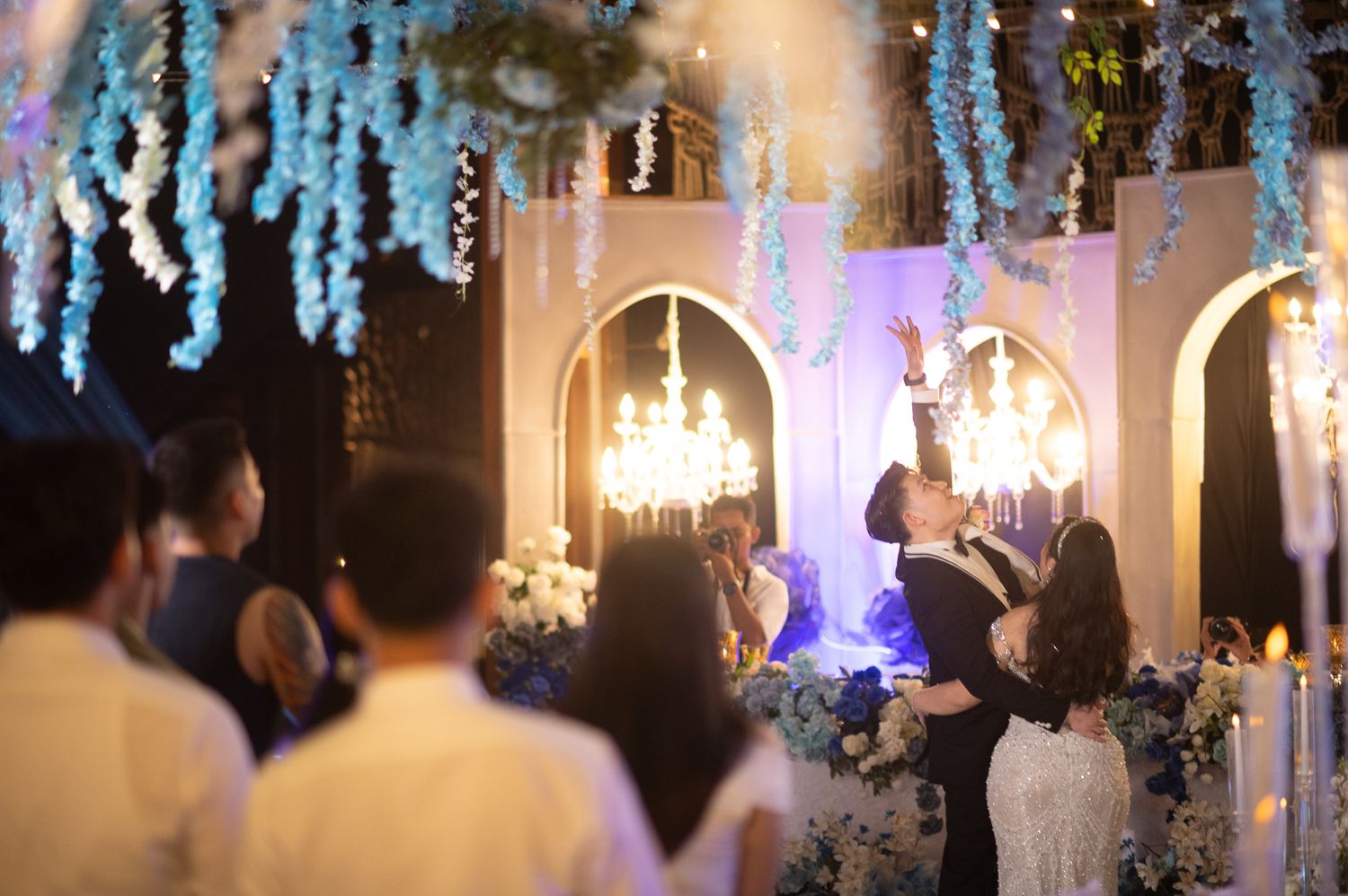 Couple sharing romantic moment at elegant wedding reception with blue floral decor and chandelier lighting.