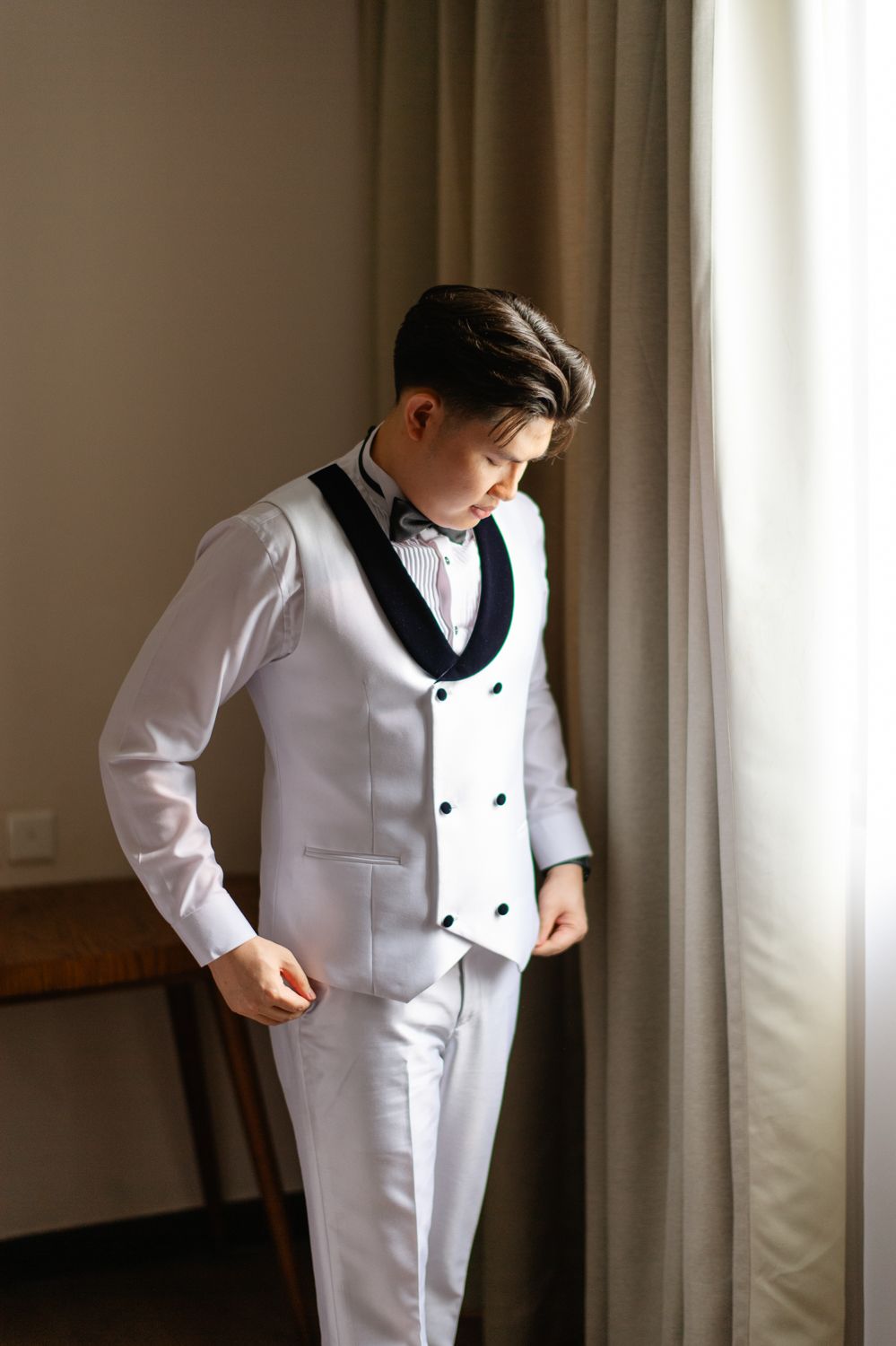 Young man in white tuxedo suit with black lapels and bow tie standing near curtained window.